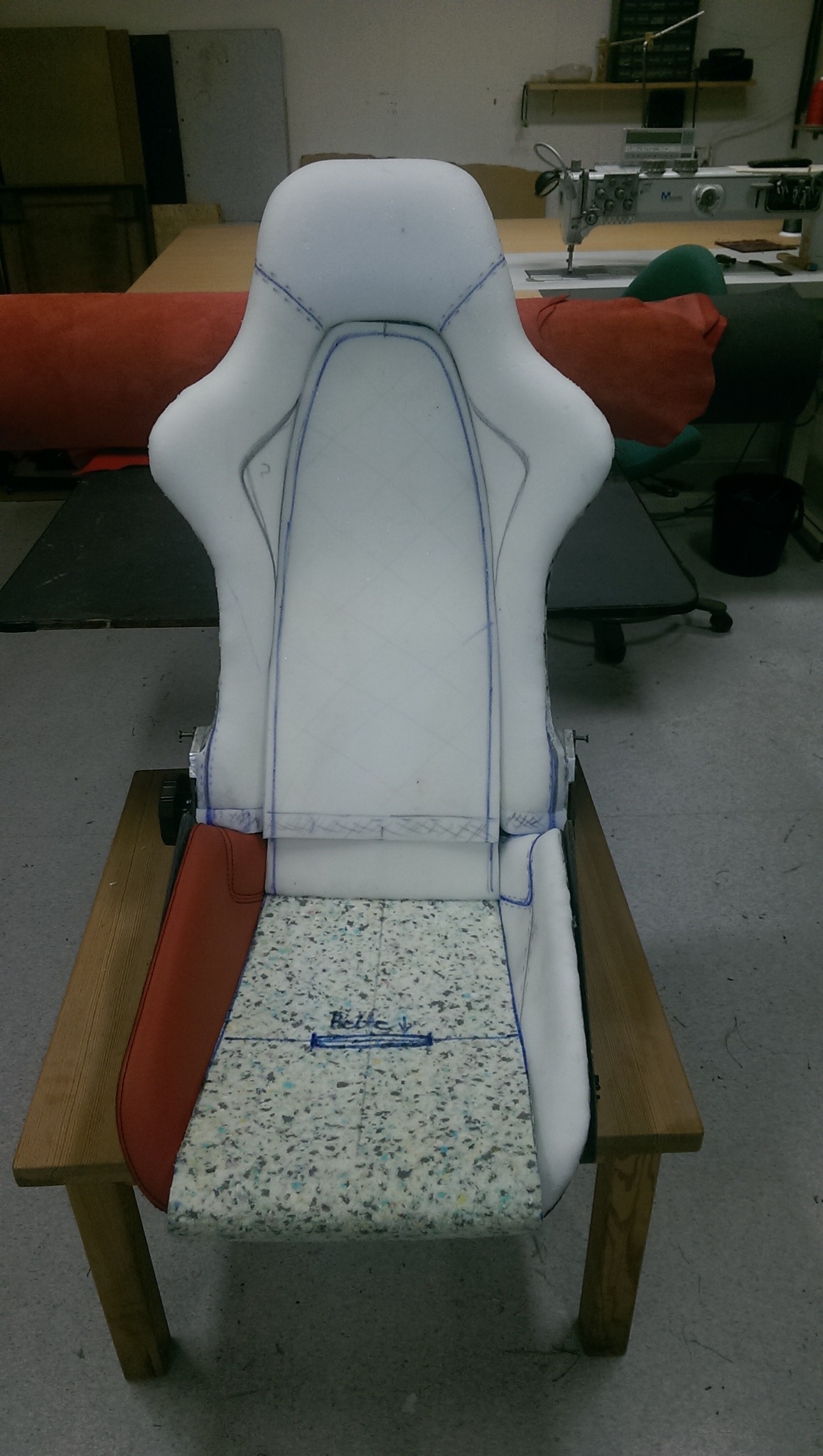 Szkielet fotela samochodowego w trakcie tapicerowania, z widocznymi warstwami pianki i zaznaczonymi liniami szwów, umieszczony na drewnianym stelażu w warsztacie.