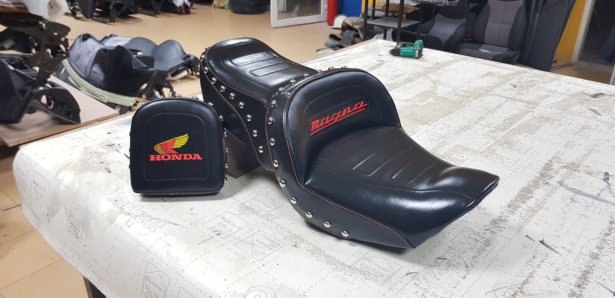Czarne, skórzane siedzenie motocyklowe z napisem Magna i mała torba z logo Honda, ozdobione srebrnymi nitami, leżą na stole w warsztacie.
