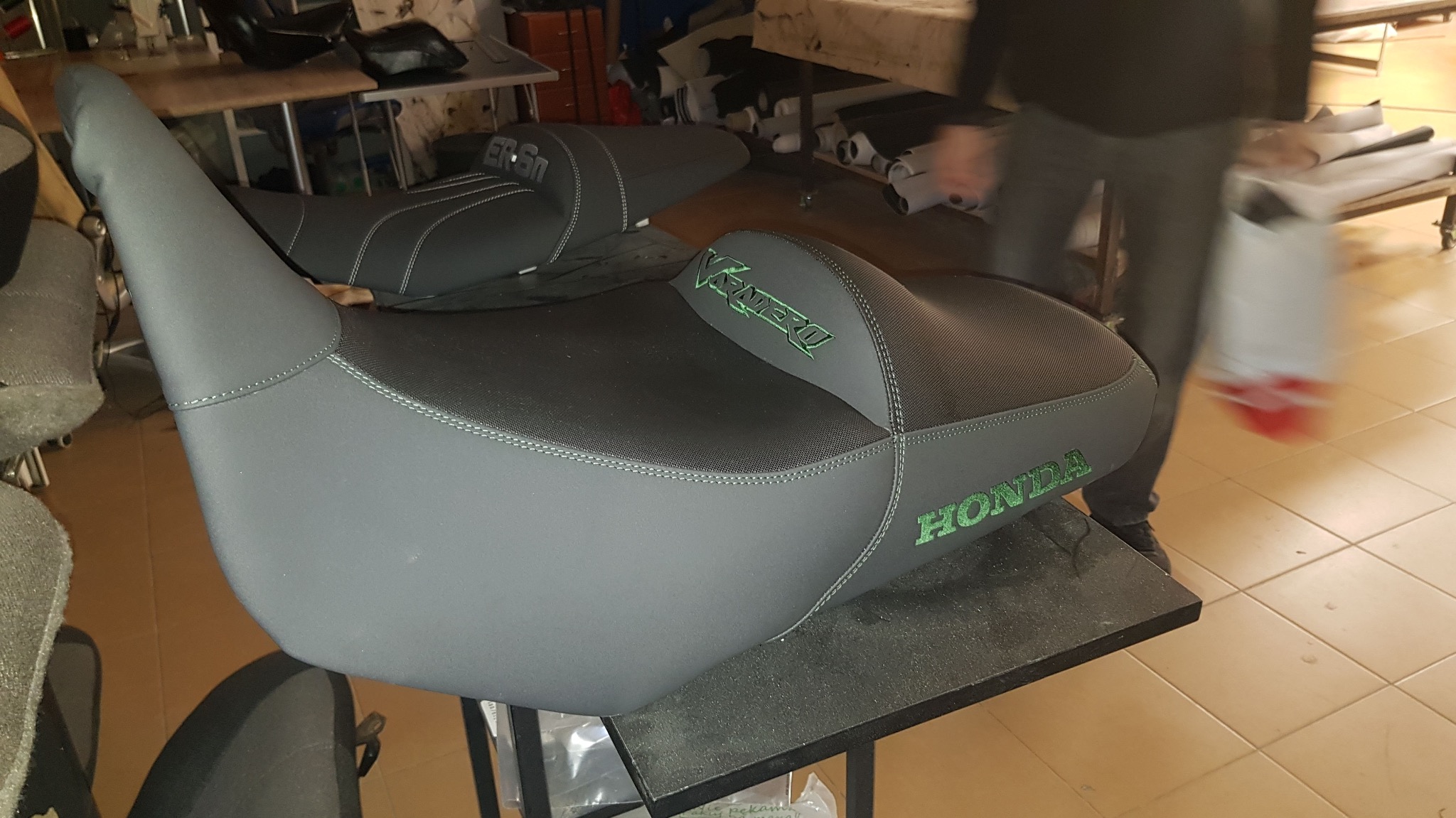 Szare, odnowione siedzenie motocyklowe Honda z zielonym napisem, widoczne szwy i inne siedzenia w tle. Tapicerka po renowacji.