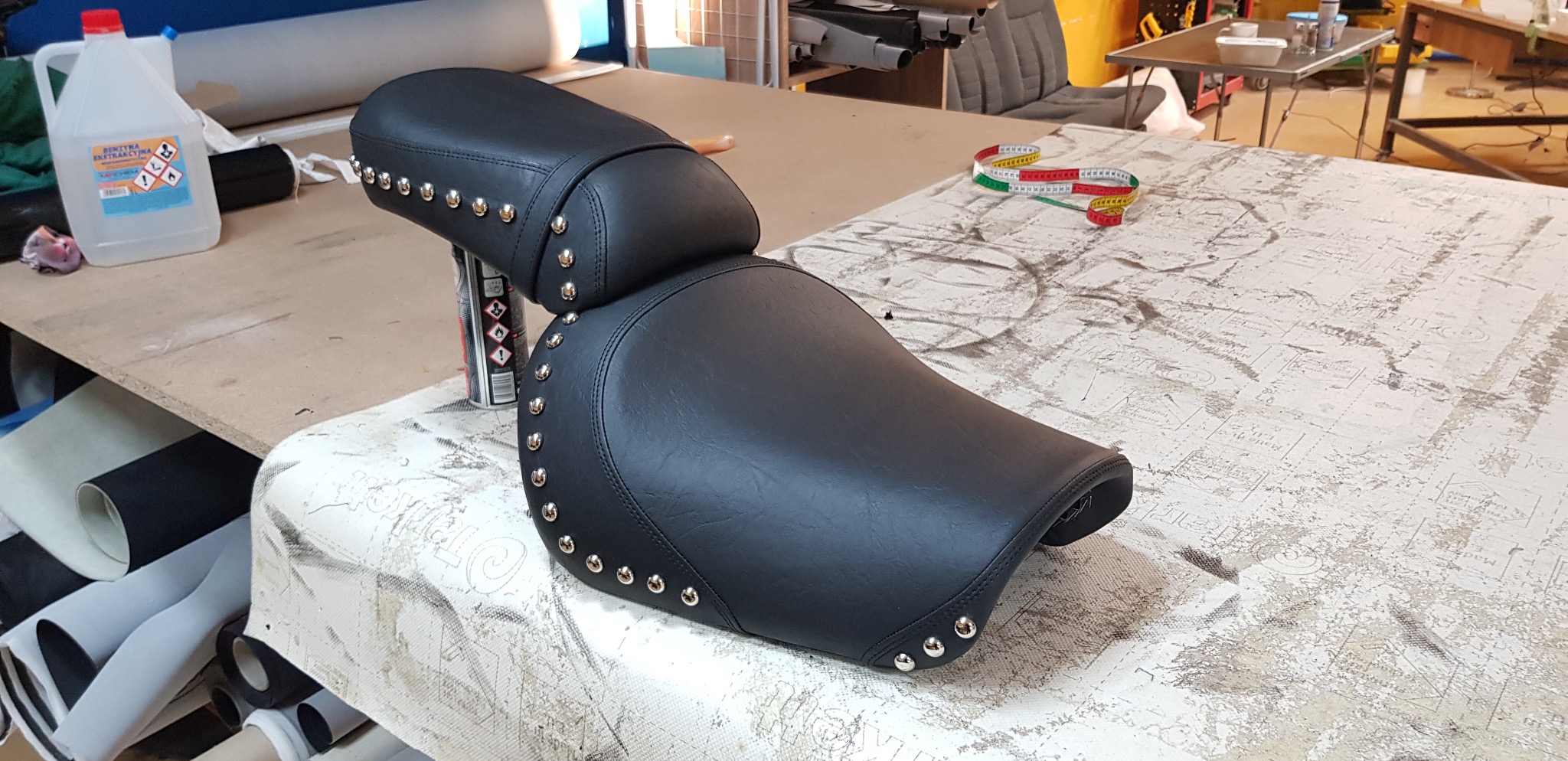 Czarne, skórzane siedzenie motocyklowe z nitami, leżące na stole w warsztacie. W tle widoczne narzędzia i materiały tapicerskie.