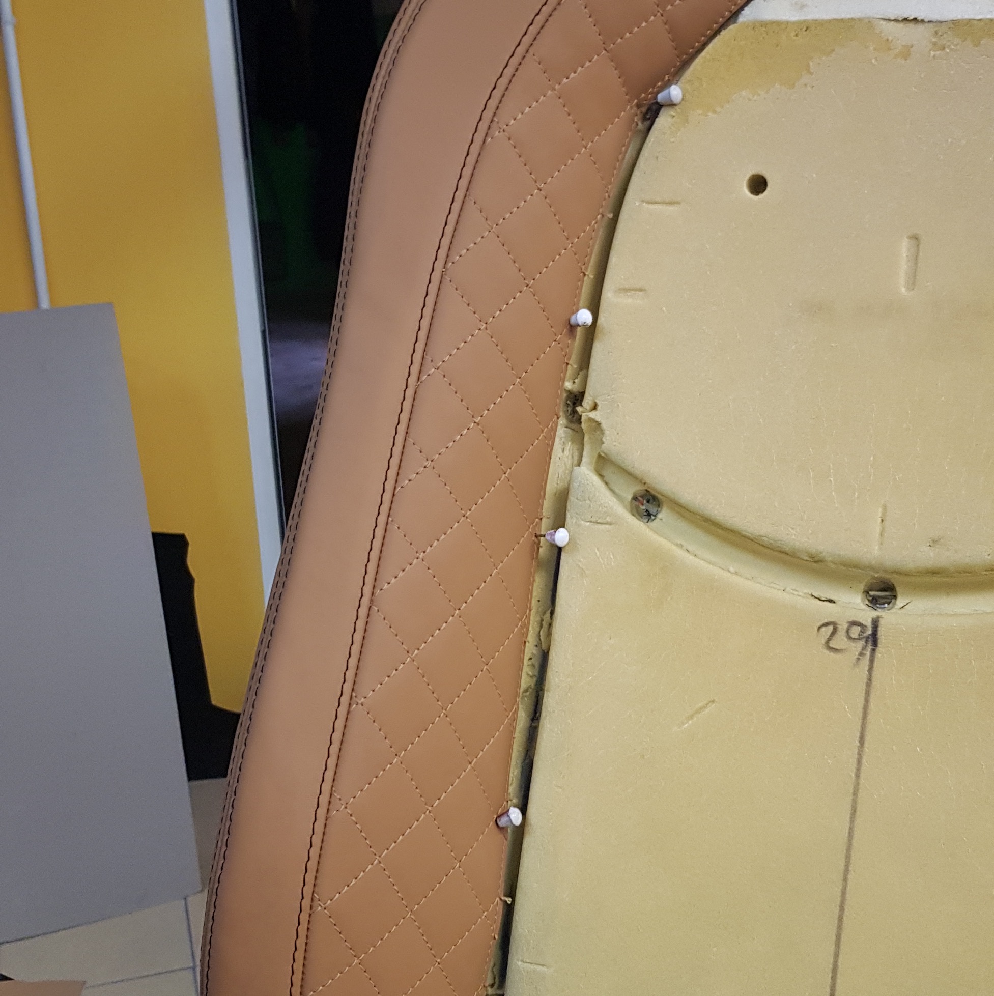 Tapicerowany element fotela samochodowego w odcieniach brązu, z widoczną strukturą rombów i piankowym wypełnieniem, przymocowany tymczasowo szpilkami.