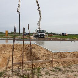 Usługi Budowlane eS-Bud Sylwester Tracz - Wylany fundament pod budowę domu z widocznym zbrojeniem, deskowaniem i pompą do betonu. Świeży beton tworzy lustrzaną powierzchnię. W tle zielone pola.
