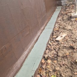 Zabezpieczona brązową masą bitumiczną ściana fundamentowa z widocznym pasem izolacji pionowej, na tle ziemi z liśćmi. Prace izolacyjne fundamentów.