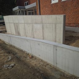 Usługi Budowlane eS-Bud Sylwester Tracz - Surowy betonowy mur oporowy w trakcie budowy, widoczny z ziemi, z ceglanym budynkiem w tle i częściowo widocznym samochodem dostawczym.