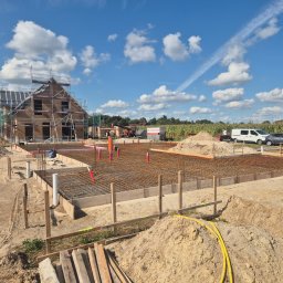 Usługi Budowlane eS-Bud Sylwester Tracz - Zbrojenie fundamentów pod budowę domu jednorodzinnego w słoneczny dzień. Widoczne szalunki, stalowe pręty, rury instalacyjne i częściowo wzniesiona konstrukcja domu.