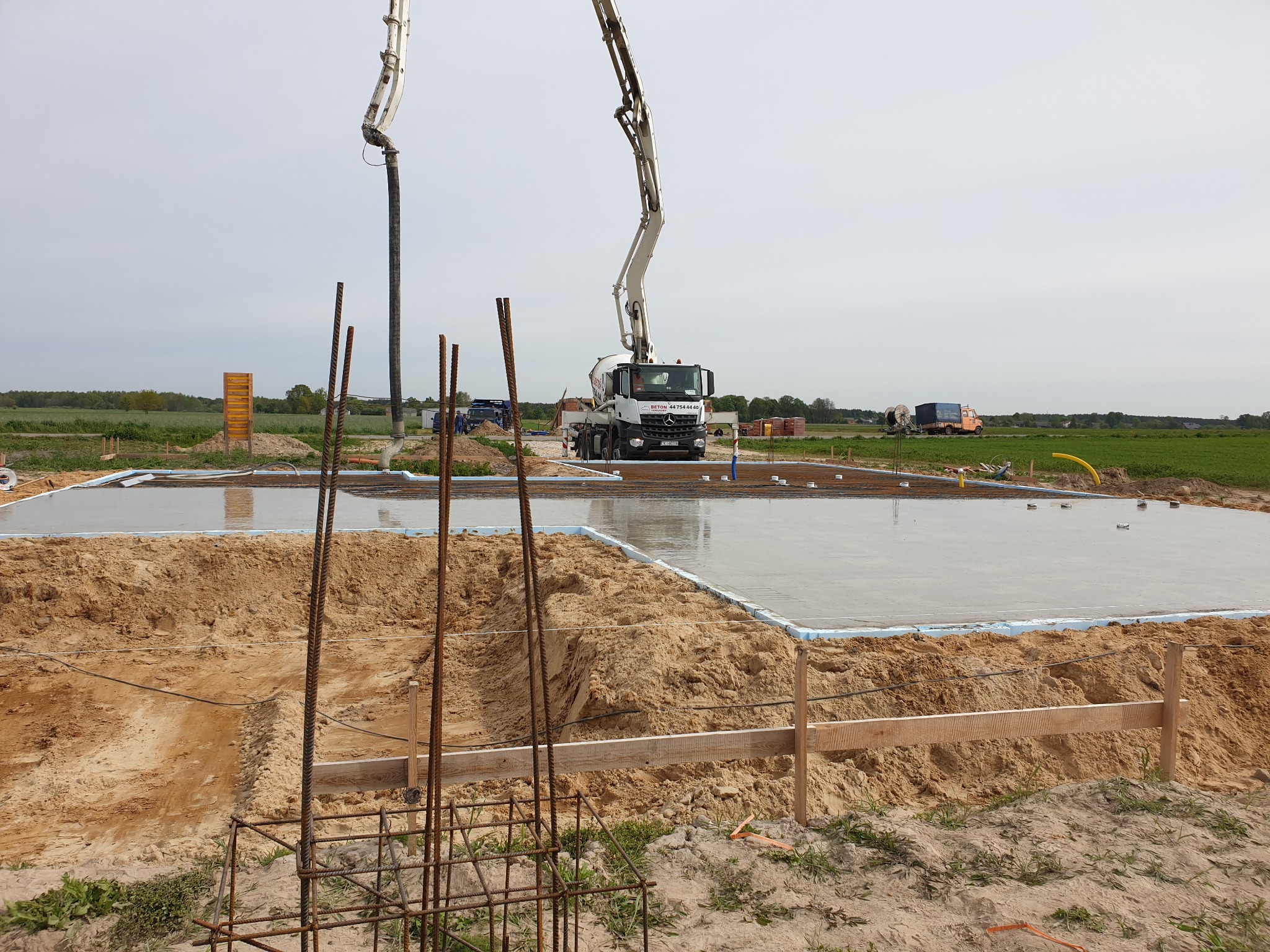 Wylany fundament pod budowę domu z widocznym zbrojeniem, deskowaniem i pompą do betonu. Świeży beton tworzy lustrzaną powierzchnię. W tle zielone pola.