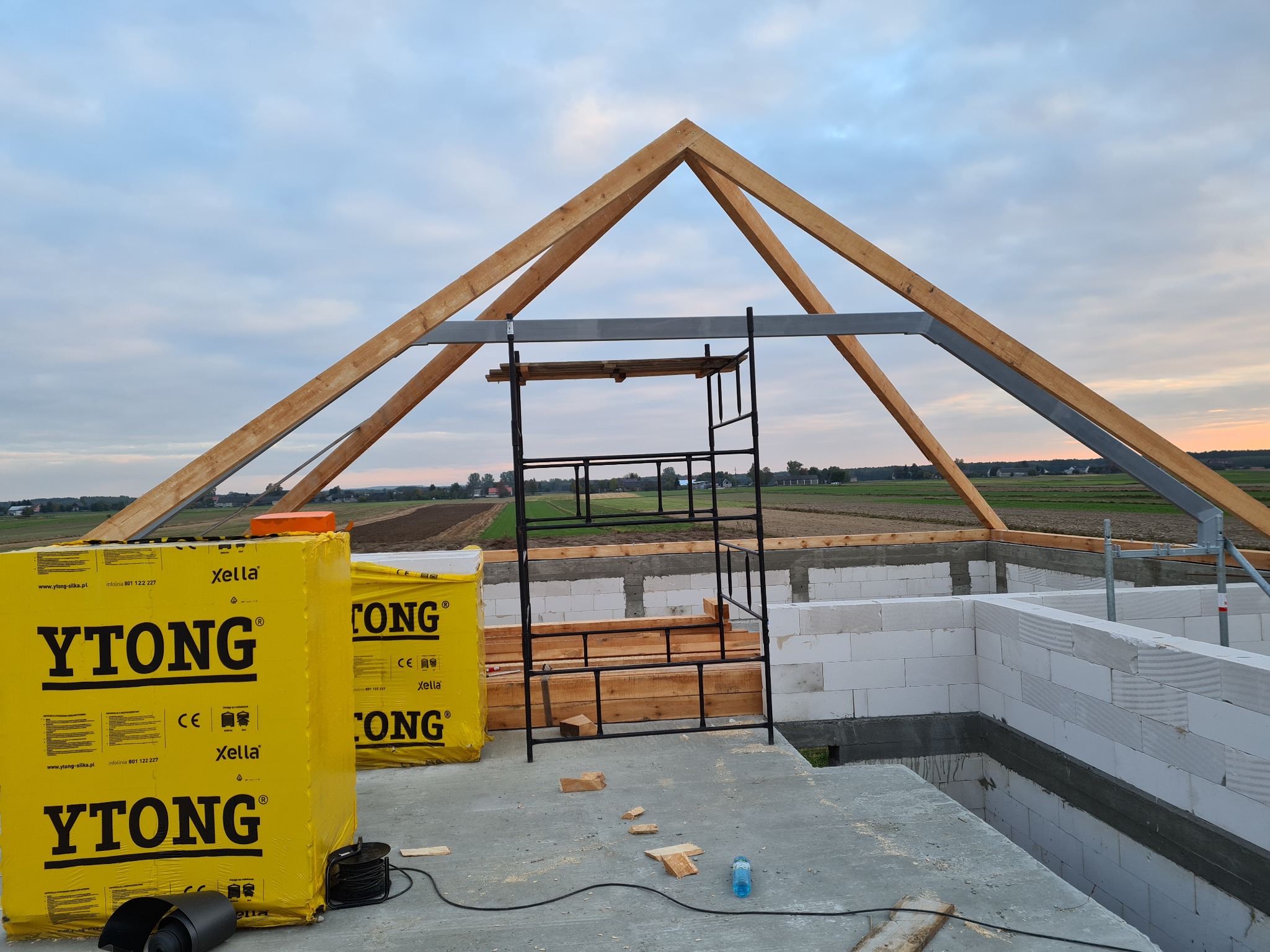 Budowa domu z YTONG: ściany z białych bloczków, drewniana konstrukcja dachu, rusztowanie. Widok na pola i zachodzące słońce w tle. Prace murarskie w toku.