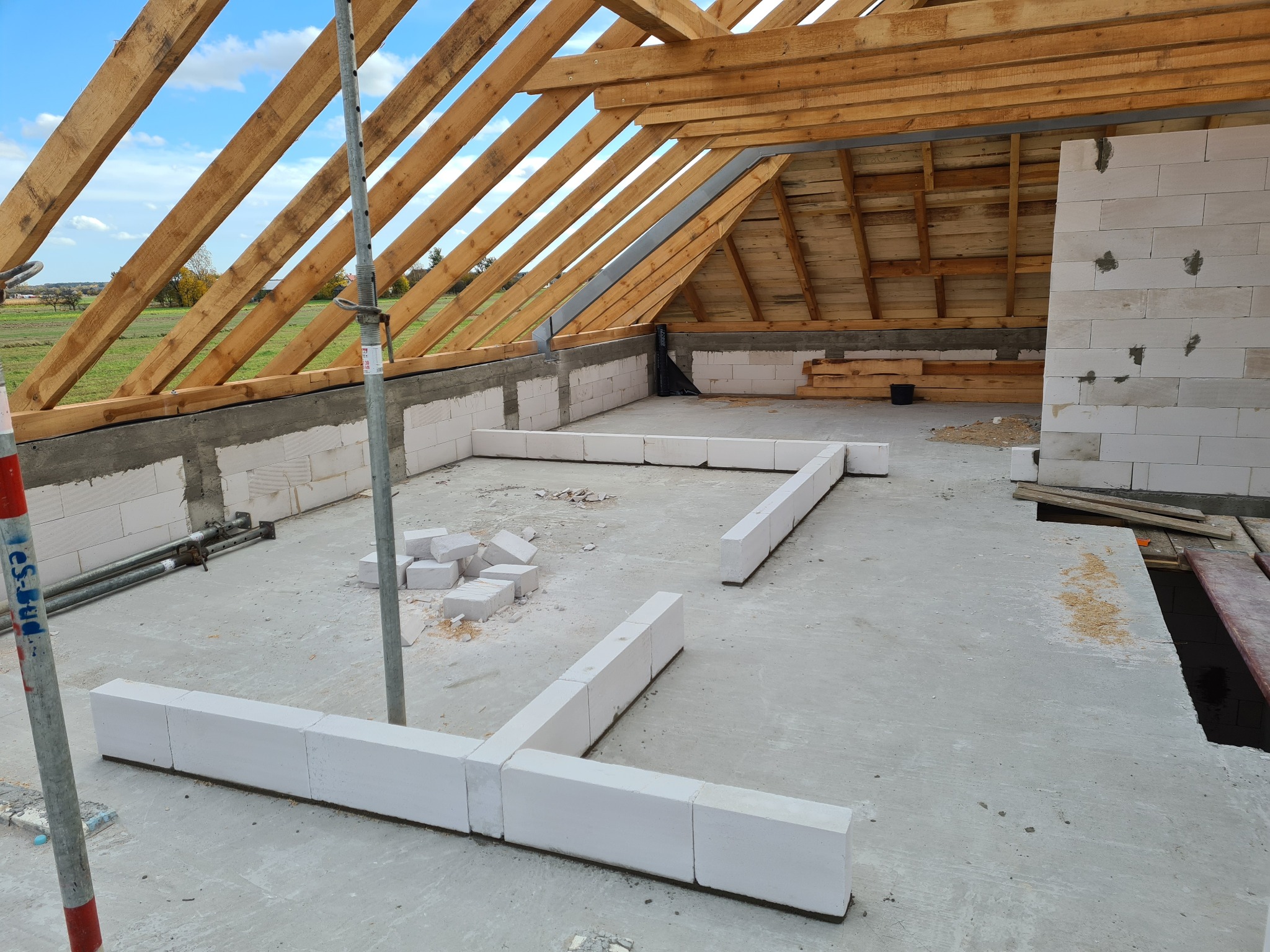 Wnętrze domu w trakcie budowy: widoczny drewniany szkielet dachu, ściany z białych bloczków, częściowo ułożone na podłodze. W tle zielone pola i błękitne niebo.