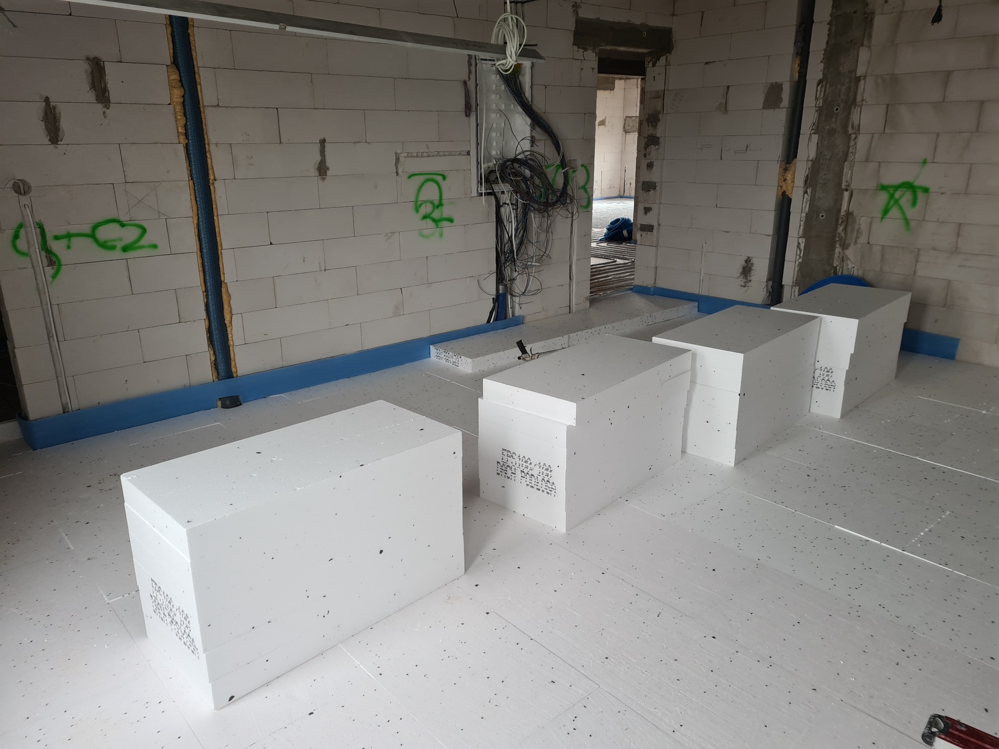 Białe płyty styropianowe ułożone na podłodze w surowym wnętrzu budynku z białych bloczków. Widoczne instalacje elektryczne i oznaczenia na ścianach.