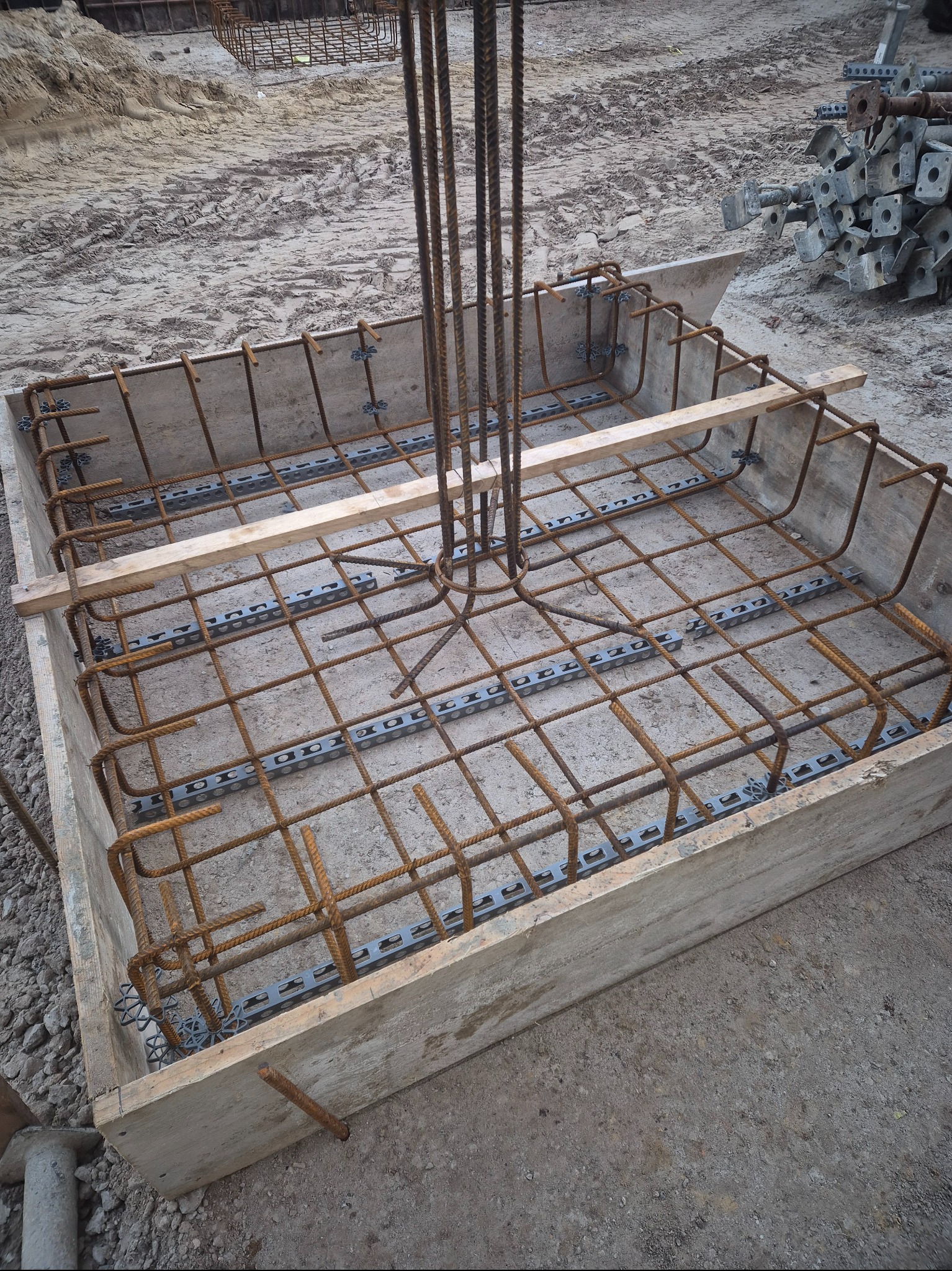Zbrojenie fundamentu w drewnianym szalunku, widoczne stalowe pręty i elementy dystansowe, przygotowane do zalania betonem. W tle ziemia i elementy konstrukcyjne.