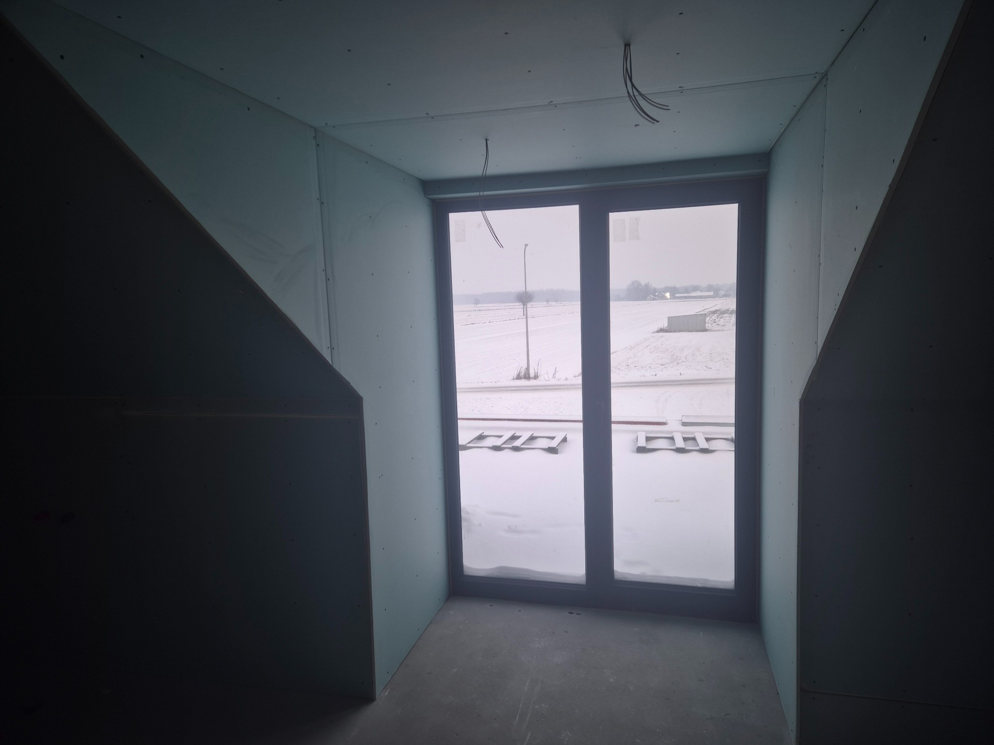Poddasze w trakcie wykończenia: ściany obłożone płytami gipsowo-kartonowymi, okno z widokiem na zimowy krajobraz. Widoczne kable elektryczne.