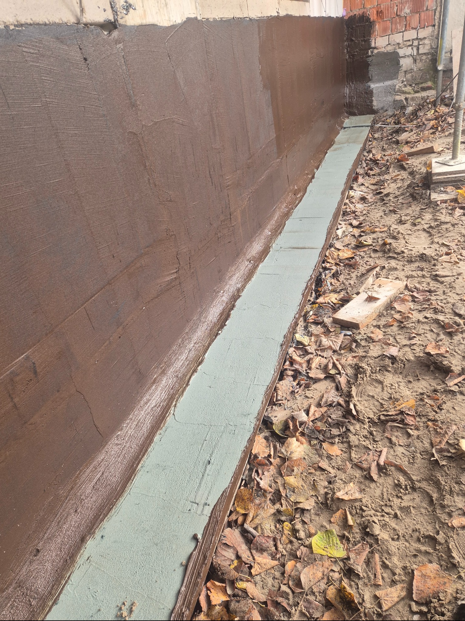 Zabezpieczona brązową masą bitumiczną ściana fundamentowa z widocznym pasem izolacji pionowej, na tle ziemi z liśćmi. Prace izolacyjne fundamentów.