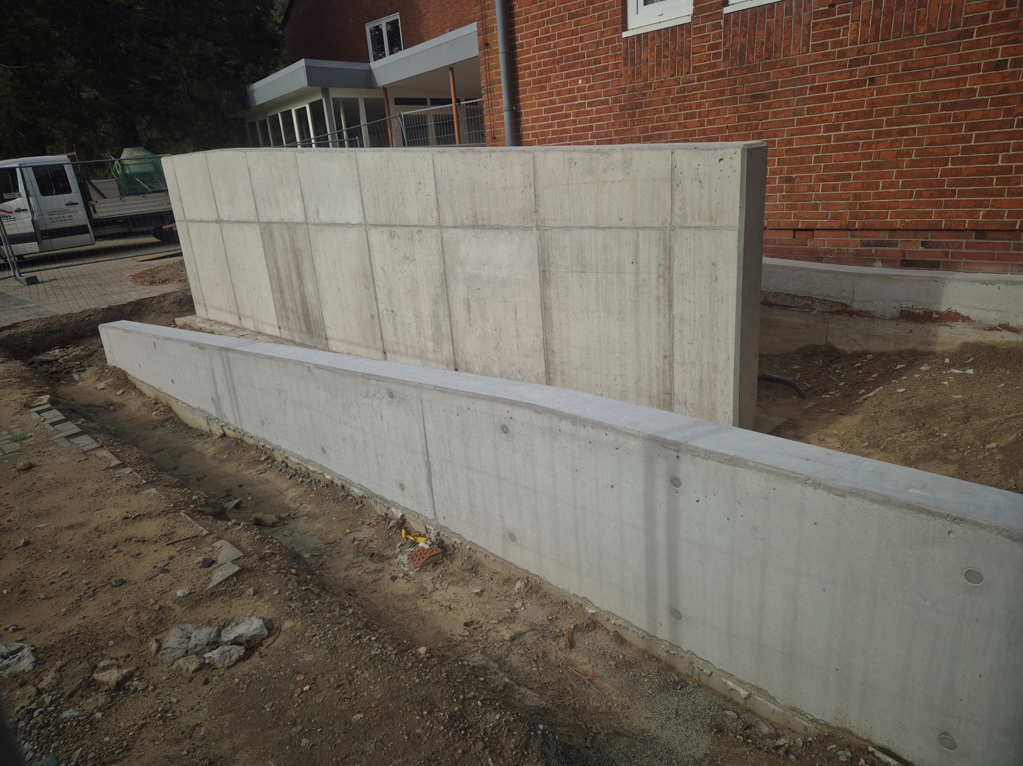 Surowy betonowy mur oporowy w trakcie budowy, widoczny z ziemi, z ceglanym budynkiem w tle i częściowo widocznym samochodem dostawczym.