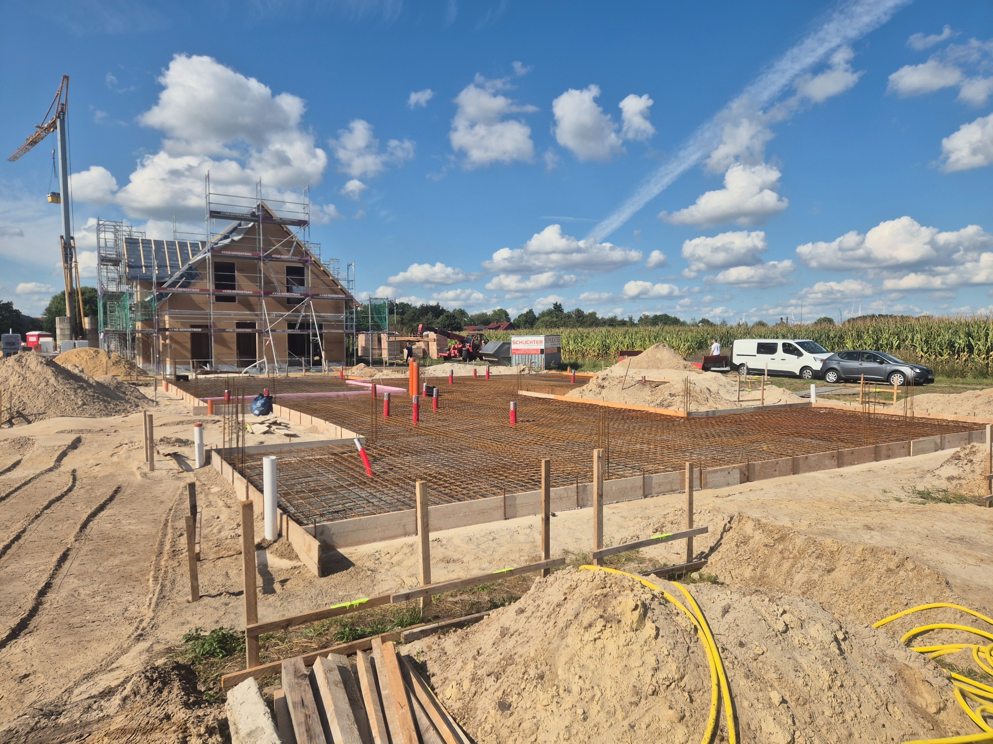 Zbrojenie fundamentów pod budowę domu jednorodzinnego w słoneczny dzień. Widoczne szalunki, stalowe pręty, rury instalacyjne i częściowo wzniesiona konstrukcja domu.
