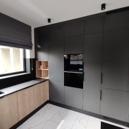 EA studio - Nowoczesna kuchnia na wymiar: czarne szafki z wbudowanym piekarnikiem, jasne drewniane blaty i szafki dolne, czarny zlew z złotą baterią, marmurowe płytki podłogowe.