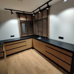 EA studio - Nowoczesna kuchnia z drewnianymi frontami szafek i czarnym blatem, oświetlona reflektorami. Widoczne drewniane żaluzje w oknie i jasna podłoga.