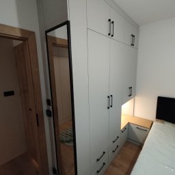 EA studio - Jasnoszara szafa wnękowa z czarnymi uchwytami, zintegrowana z szafką nocną i lustrem w czarnej ramie, odbijającym wnętrze pokoju. Widok na łóżko z materacem.