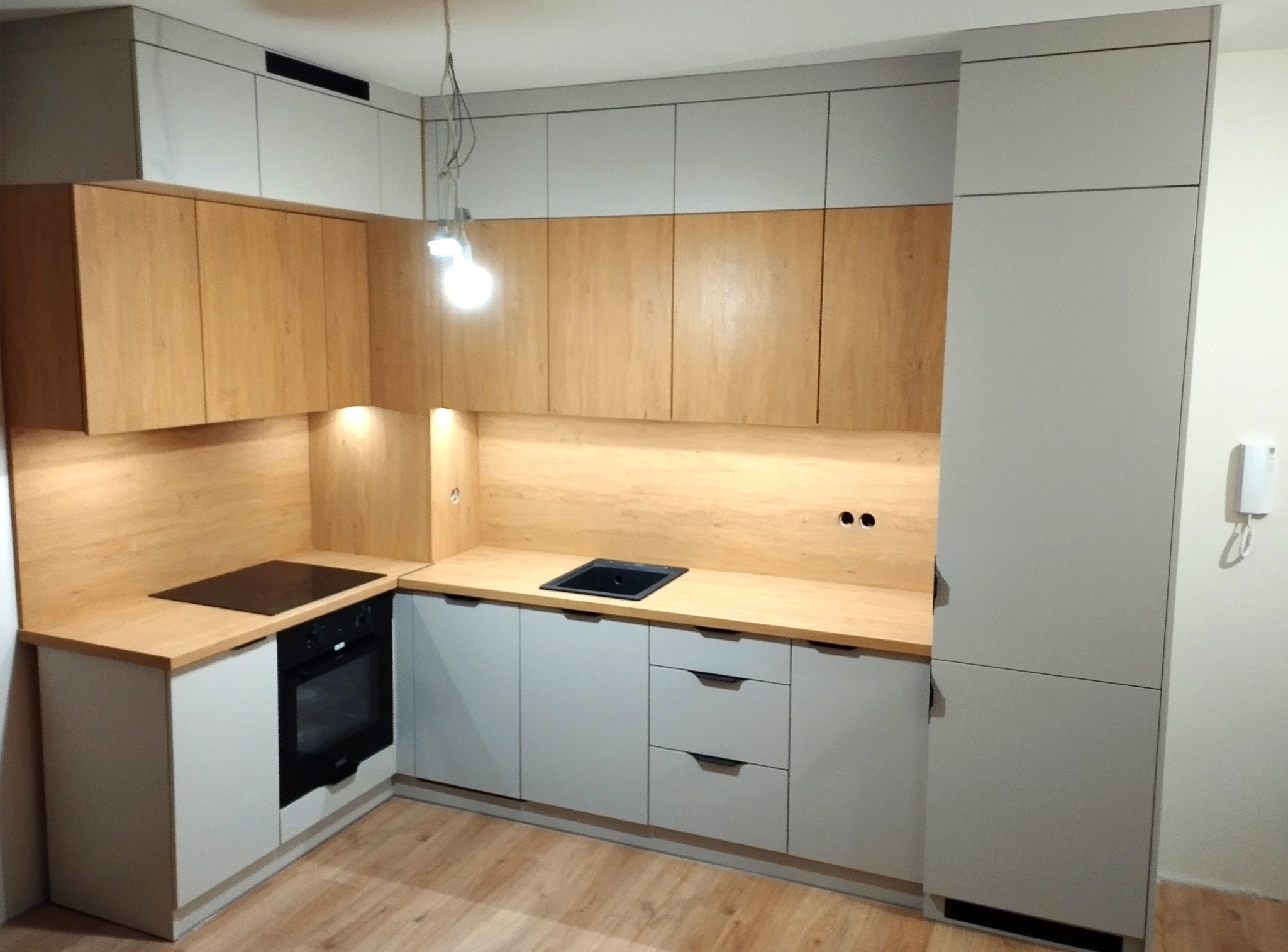 Jasna kuchnia z drewnianymi akcentami: szare szafki dolne i górne, drewniany blat, płyta indukcyjna i piekarnik. Minimalistyczny design z oświetleniem punktowym.