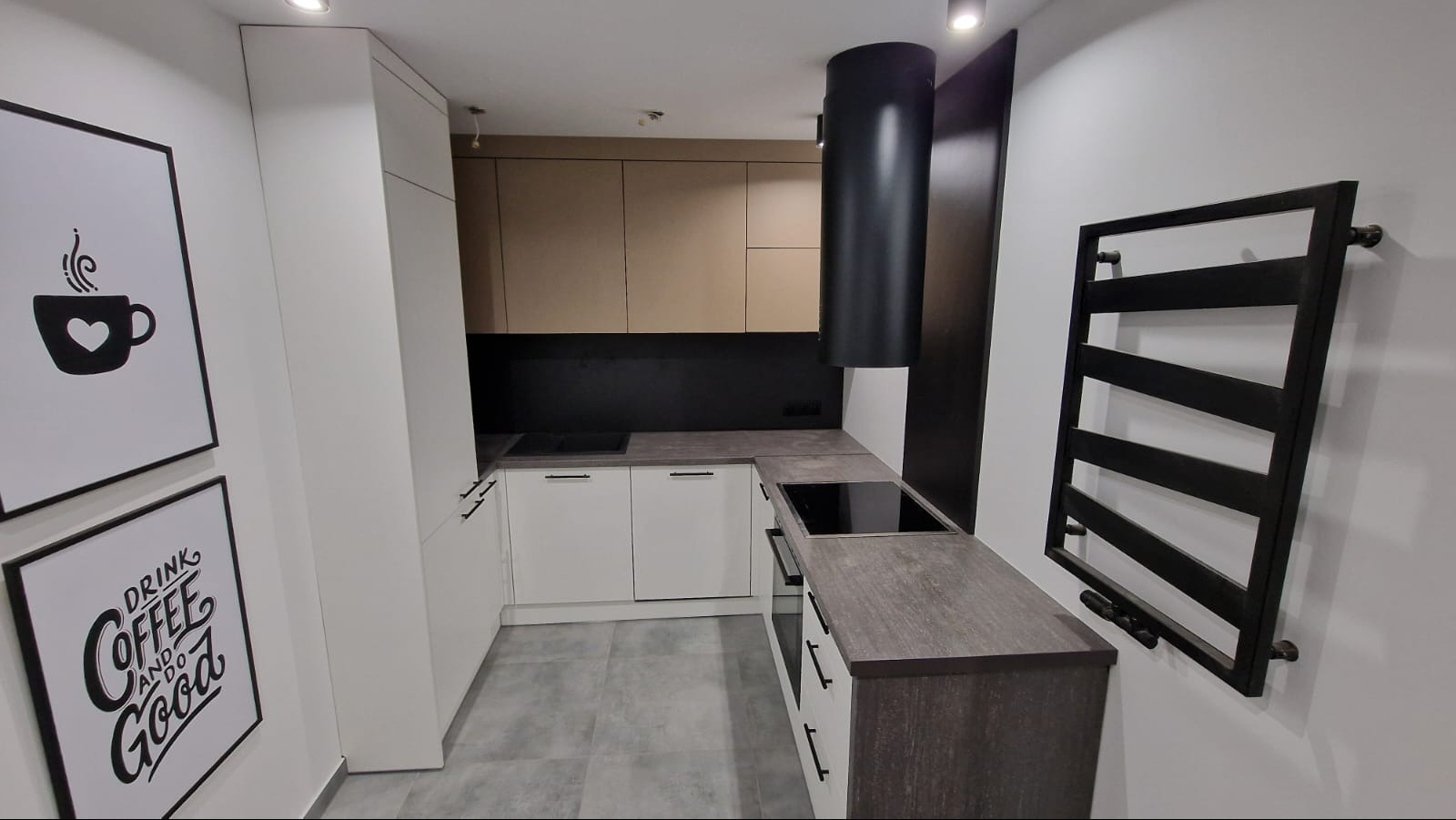Nowoczesna kuchnia w kształcie litery L z białymi i beżowymi szafkami, czarnym okapem i grzejnikiem drabinkowym. Na ścianie grafiki z motywem kawy.