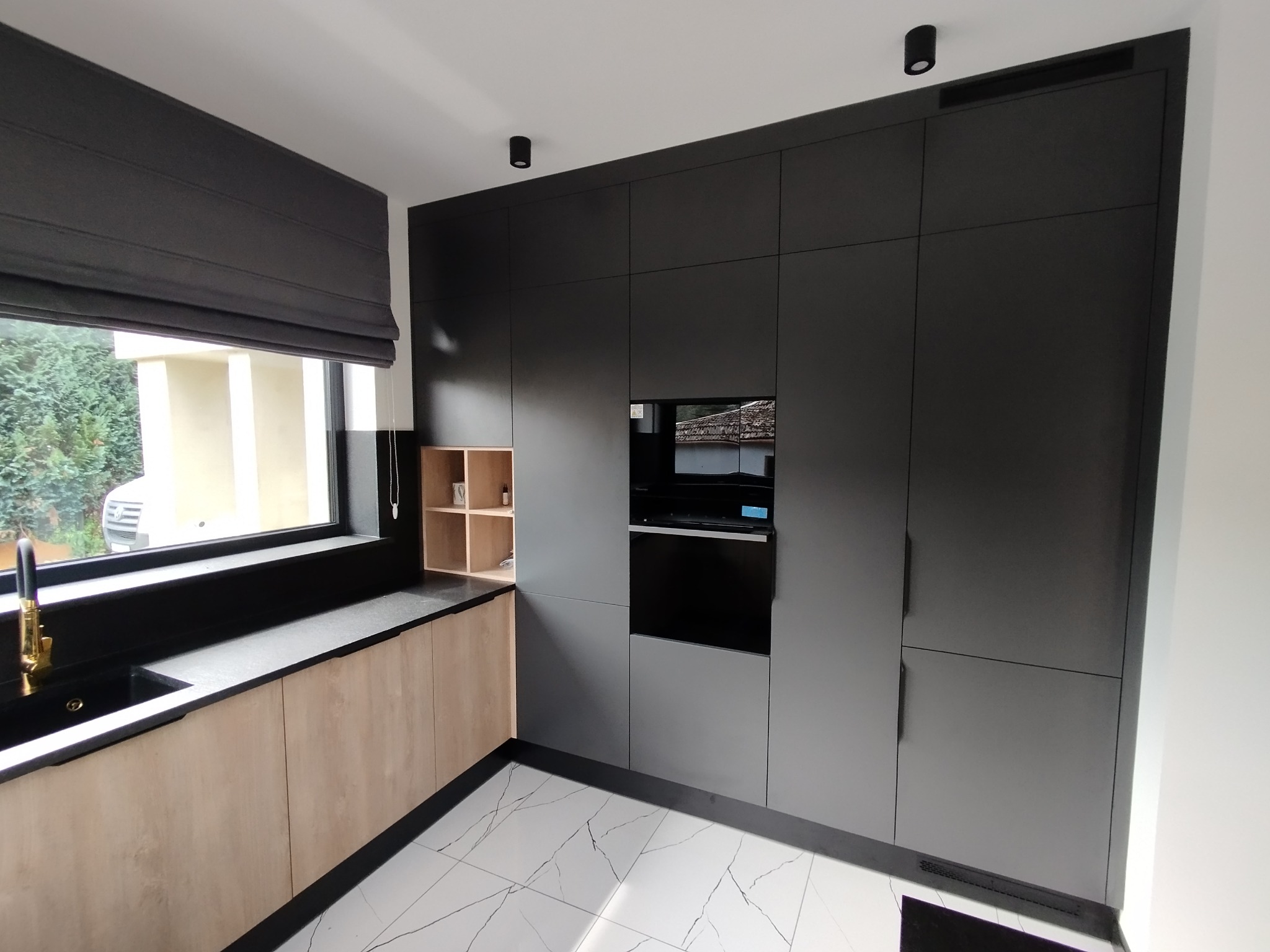 Nowoczesna kuchnia na wymiar: czarne szafki z wbudowanym piekarnikiem, jasne drewniane blaty i szafki dolne, czarny zlew z złotą baterią, marmurowe płytki podłogowe.