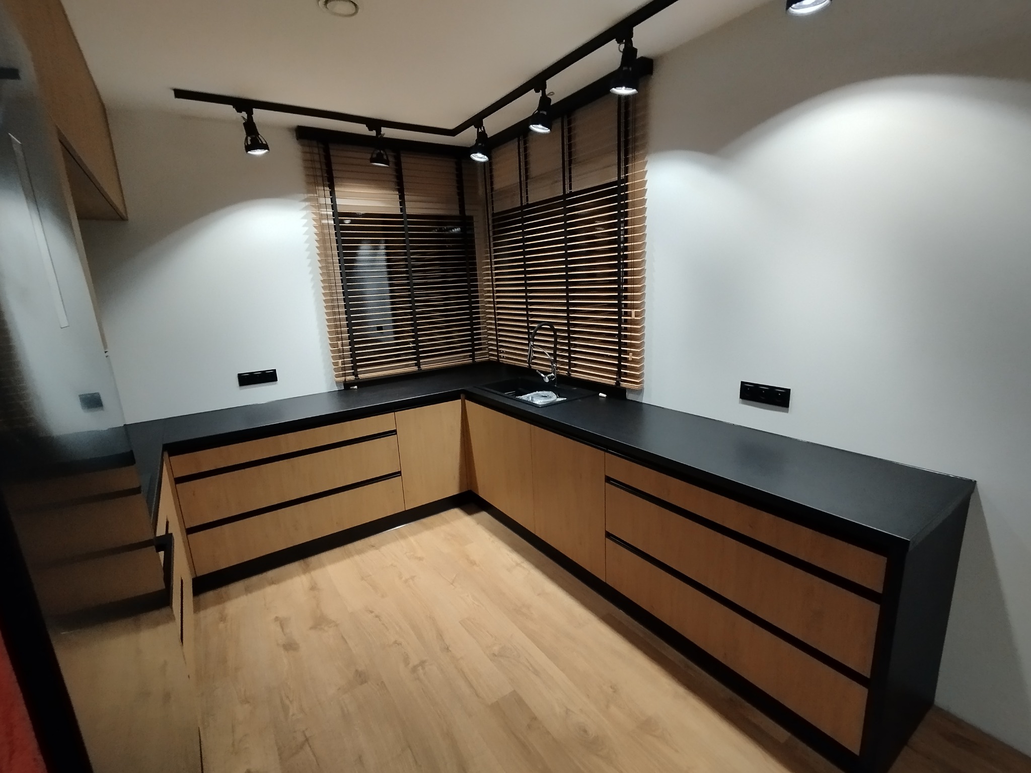 Nowoczesna kuchnia z drewnianymi frontami szafek i czarnym blatem, oświetlona reflektorami. Widoczne drewniane żaluzje w oknie i jasna podłoga.