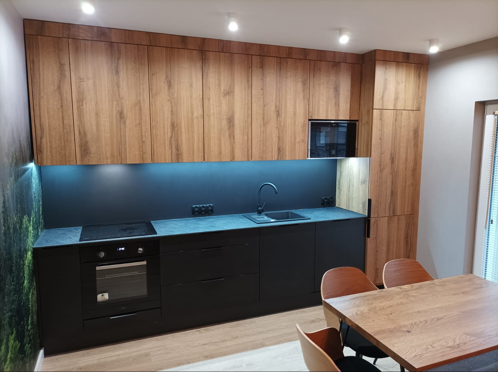 Nowoczesna kuchnia z drewnianymi szafkami górnymi i czarnymi dolnymi, oświetleniem LED pod szafkami i blatem z ciemnego kamienia, widok na stół z krzesłami.