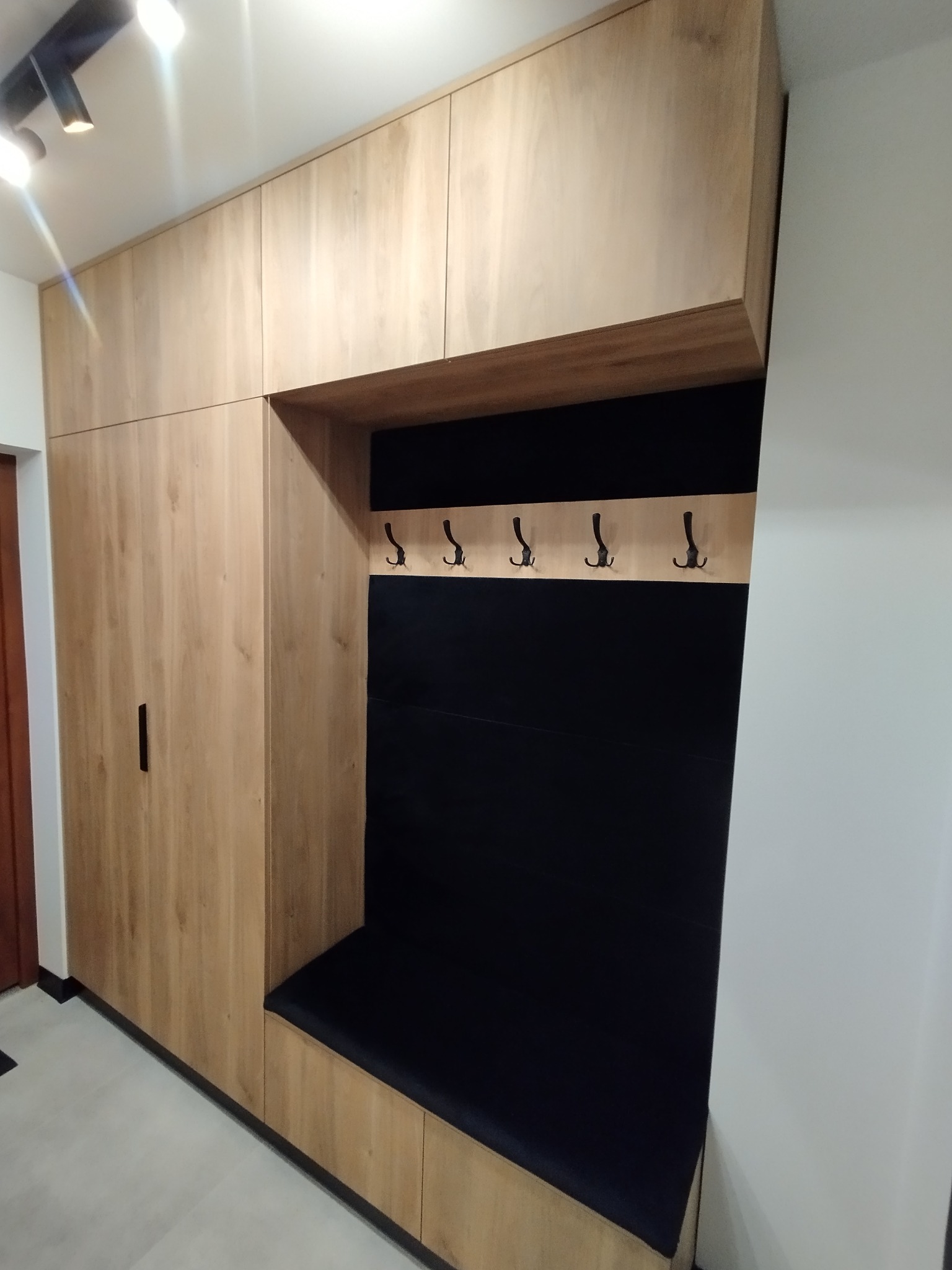 Drewniana zabudowa przedpokoju z siedziskiem i wieszakami, oświetlona punktowo. Fronty szafek z wyraźnym rysunkiem słojów. Czarna tapicerka siedziska i panelu wnęki.