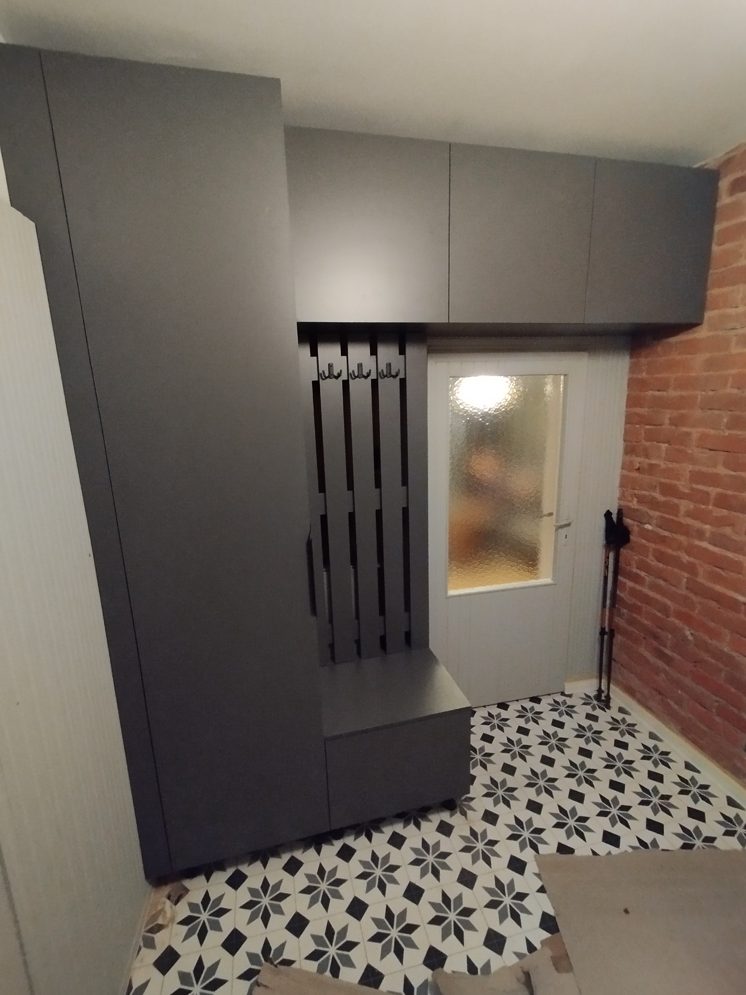 Szara zabudowa przedpokoju z siedziskiem i wieszakami, szafki wiszące, wzorzysta podłoga, fragment ceglanej ściany. Nowoczesny design, funkcjonalne rozwiązanie do przechowywania.