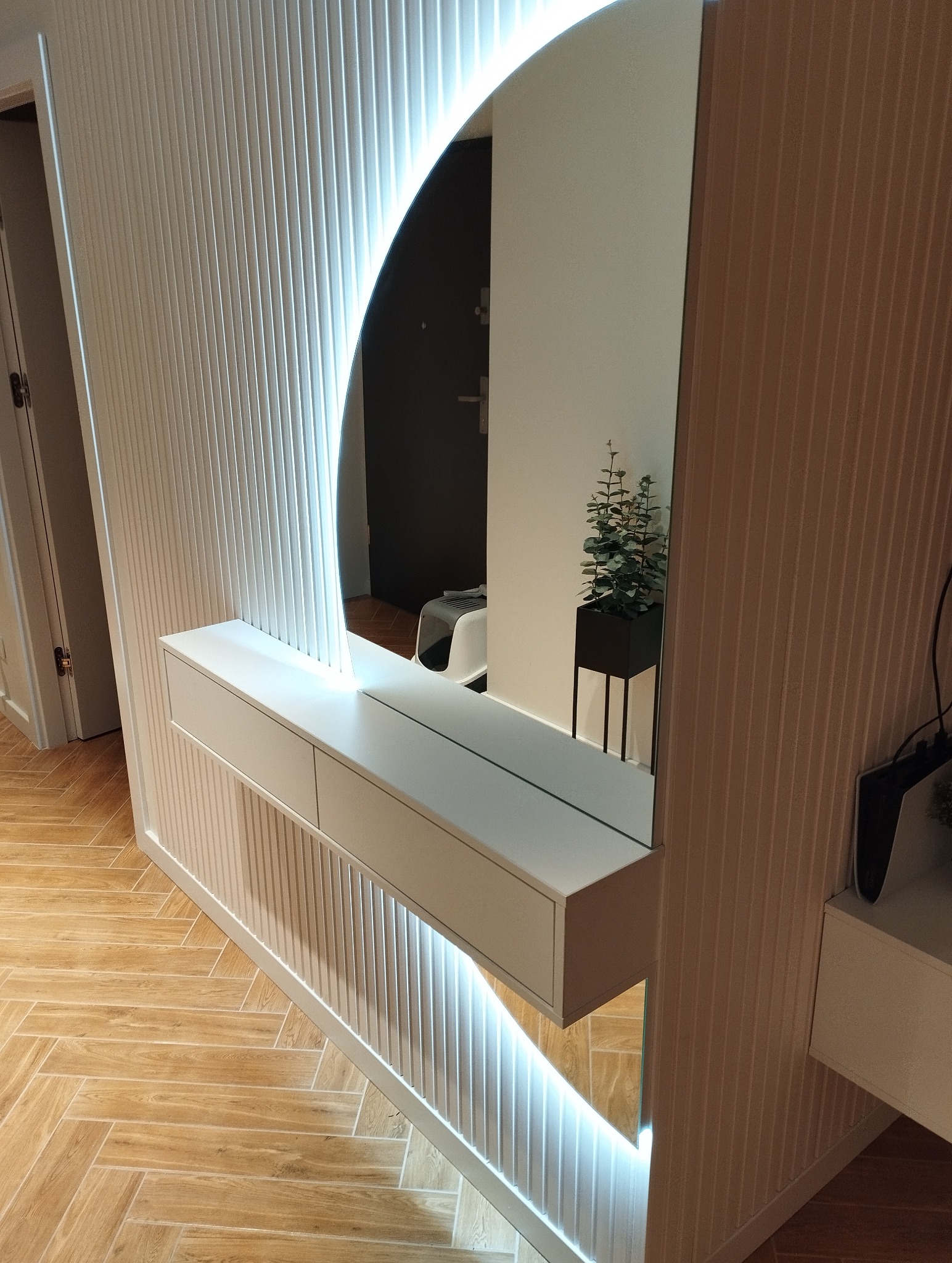 Nowoczesna zabudowa przedpokoju z lustrem w kształcie łuku, podświetlona LED, z białą konsolą i dekoracyjnymi lamelami ściennymi. Parkiet w jodełkę na podłodze.