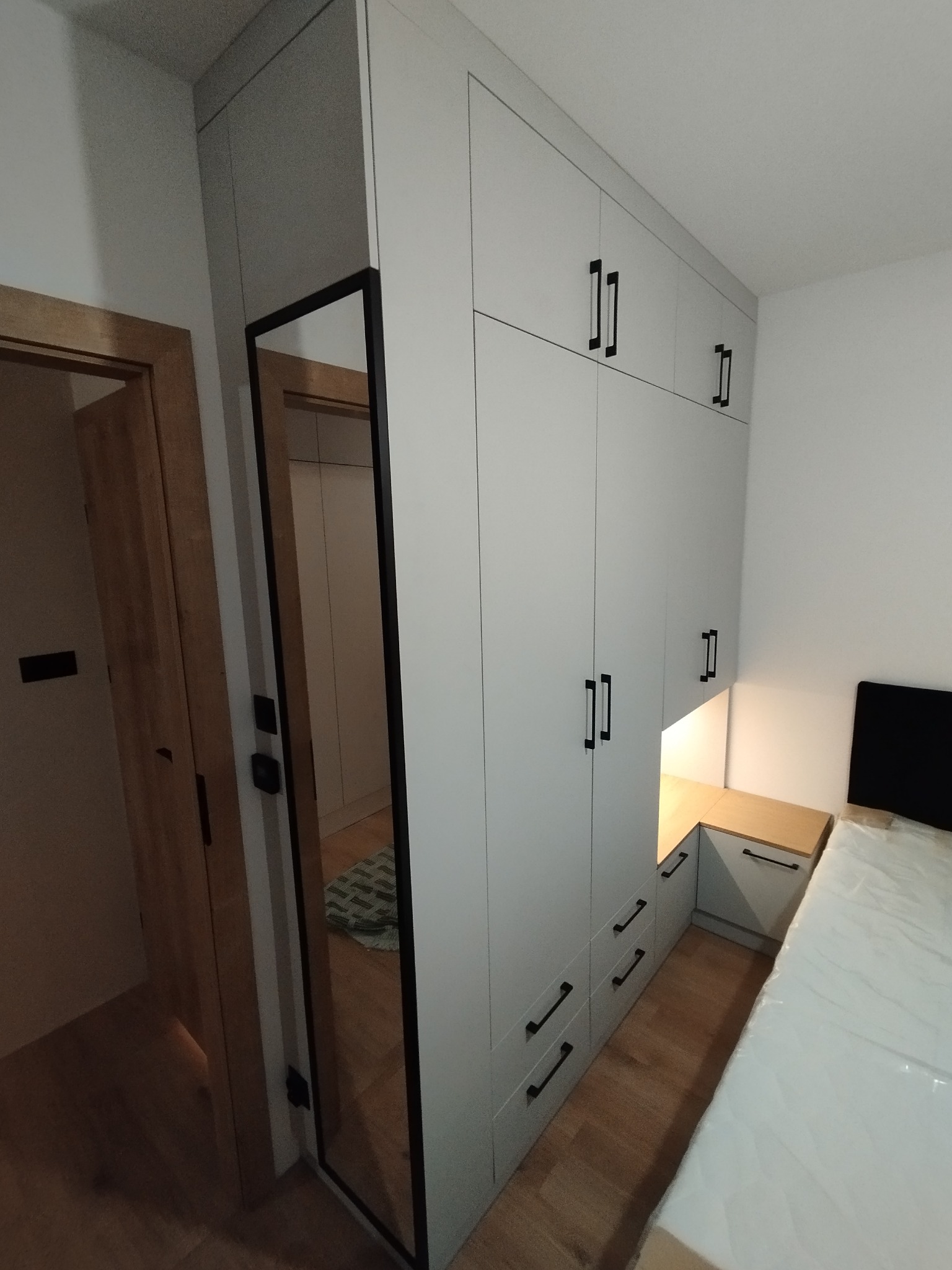 Jasnoszara szafa wnękowa z czarnymi uchwytami, zintegrowana z szafką nocną i lustrem w czarnej ramie, odbijającym wnętrze pokoju. Widok na łóżko z materacem.