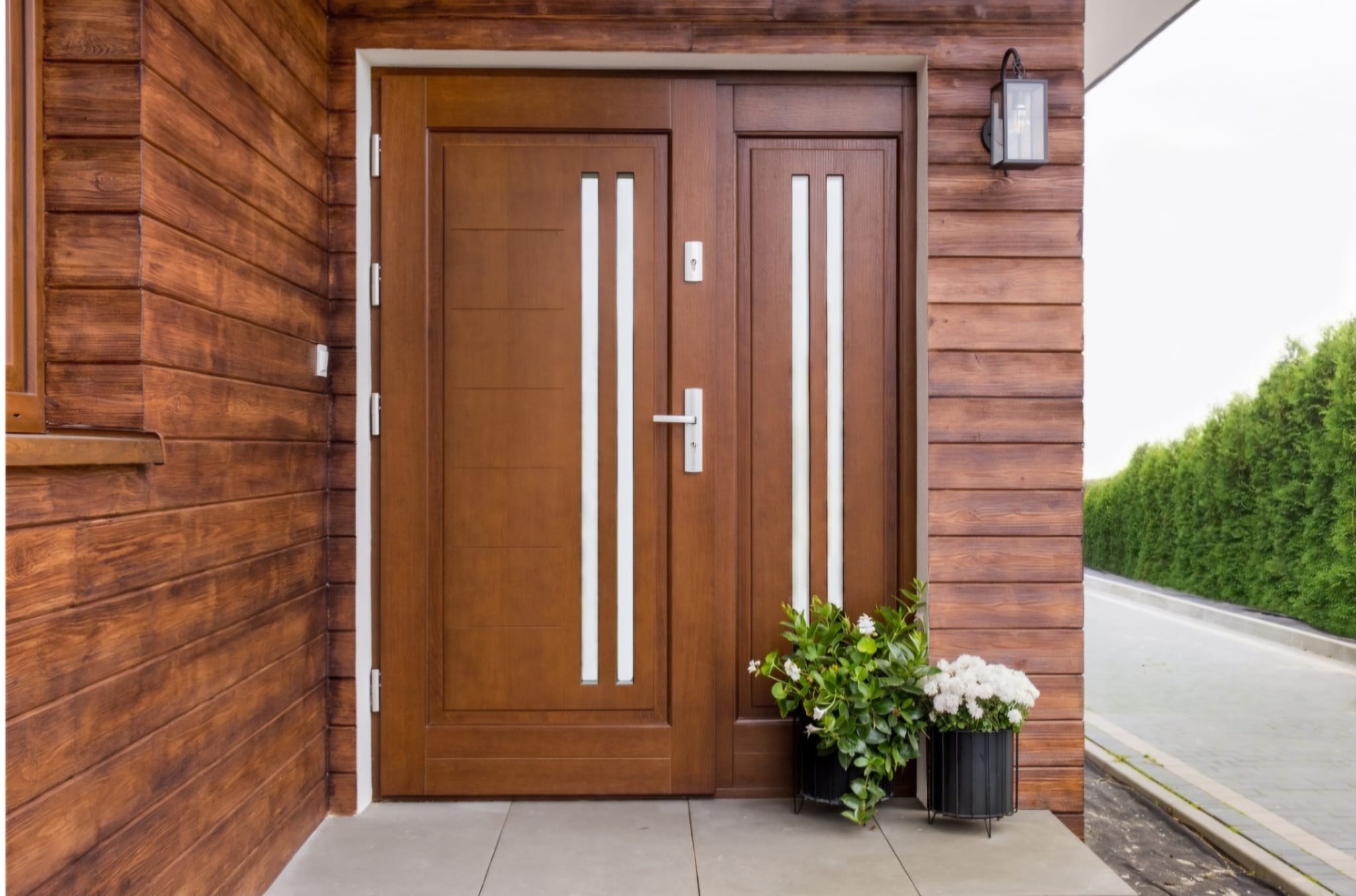 Brązowe drzwi wejściowe z przeszkleniami, obok donice z kwiatami. Elewacja budynku z drewnianą okładziną. Oświetlenie zewnętrzne.