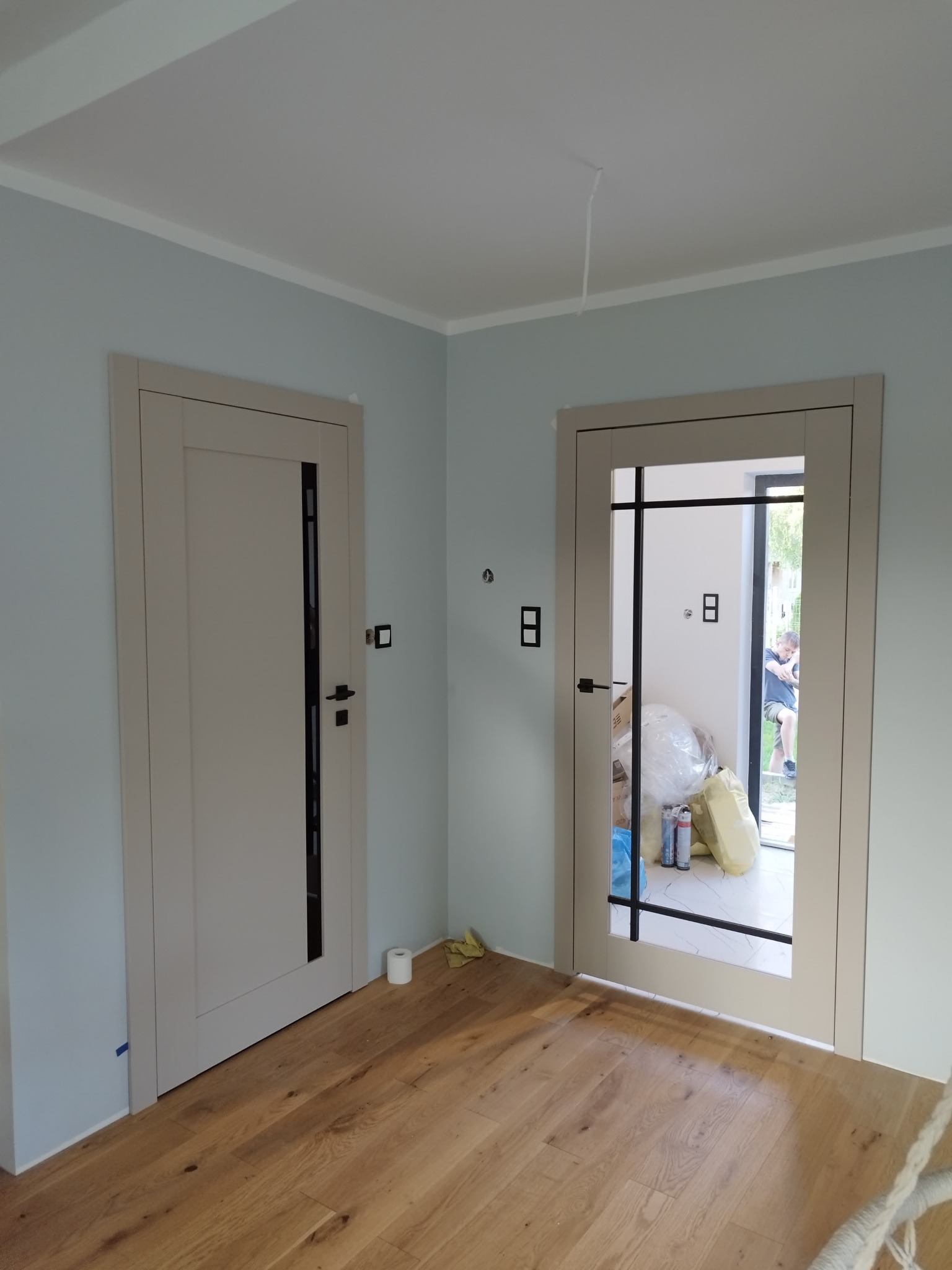 Dwa zamontowane, beżowe drzwi wewnętrzne z czarnymi klamkami i elementami dekoracyjnymi, widoczne na tle jasnych ścian i drewnianej podłogi.