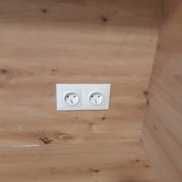 Koliber Studio - Dwa białe gniazdka elektryczne zamontowane w drewnianej płycie meblowej o jasnym, naturalnym odcieniu. Widoczne słoje drewna i subtelne detale wykończenia.