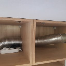 Koliber Studio - Wnętrze szafki z zamontowanym, elastycznym przewodem wentylacyjnym. Widoczna listwa zasilająca. Jasne drewno i białe elementy tworzą minimalistyczny design.