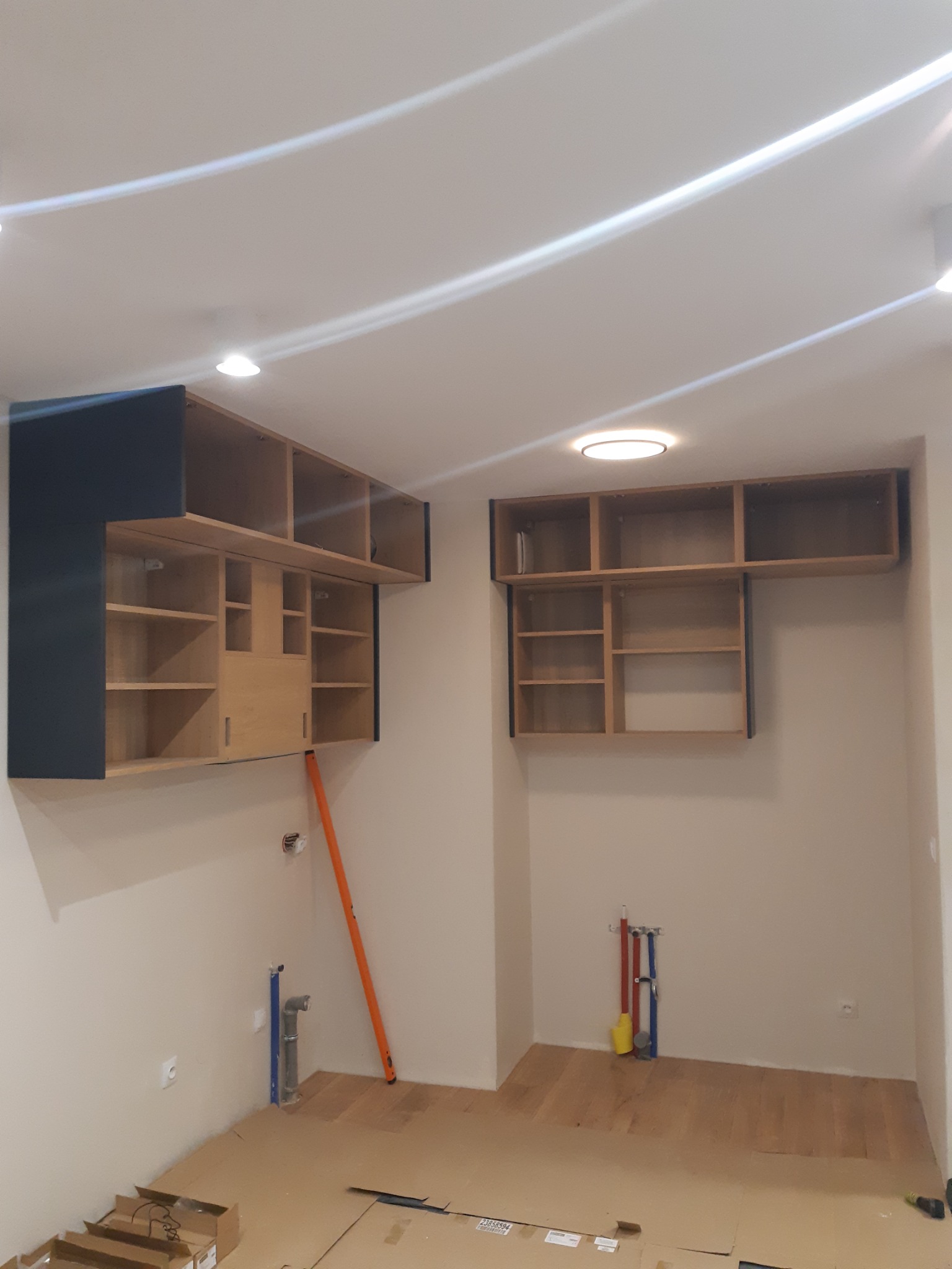 Montaż wiszących szafek kuchennych z drewnianymi półkami i ciemnymi obramowaniami, poziomica oparta o ścianę, widoczne instalacje wodne, podłoga zabezpieczona kartonem.