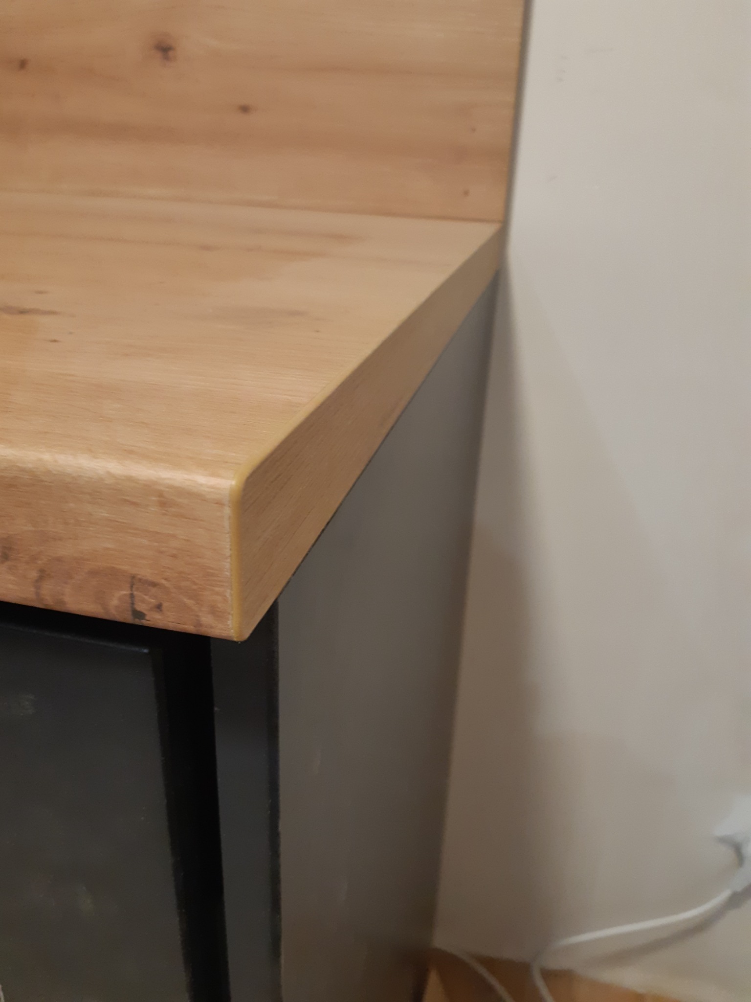 Fragment blatu z drewna o ciepłym odcieniu, zamontowanego na szafce w kolorze grafitowym, przylegającego do jasnej ściany. Widoczne detale wykończenia krawędzi blatu.