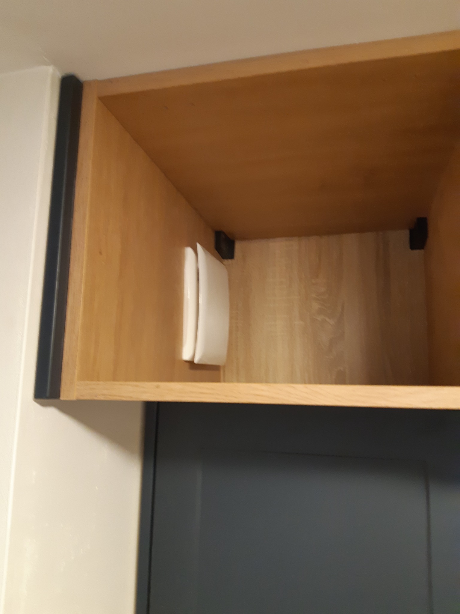 Wnętrze szafki kuchennej z jasnego drewna, z zamontowanym białym, podwójnym włącznikiem światła. Widoczna szara frontowa część szafki i fragment białej ściany.