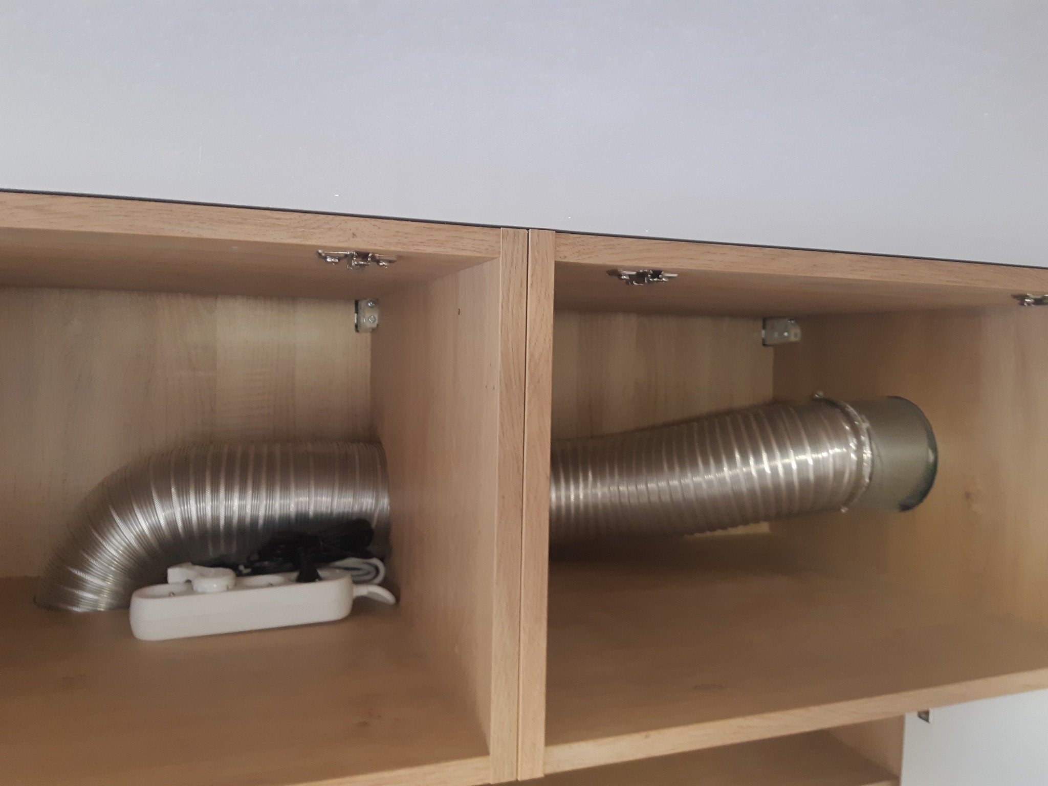 Wnętrze szafki z zamontowanym, elastycznym przewodem wentylacyjnym. Widoczna listwa zasilająca. Jasne drewno i białe elementy tworzą minimalistyczny design.