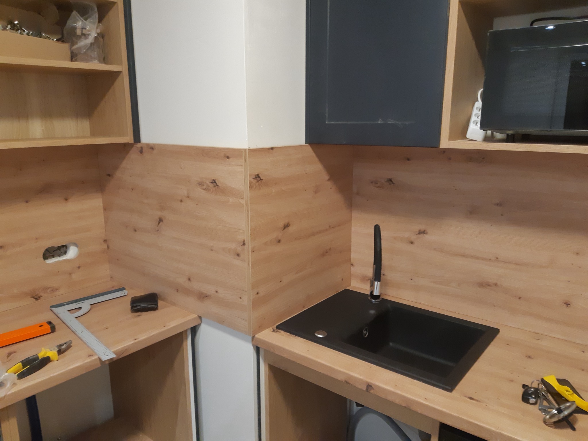 Częściowo wykończona kuchnia z drewnianymi panelami, czarnym zlewem i widocznymi narzędziami, sugerująca etap montażu lub wykończenia.