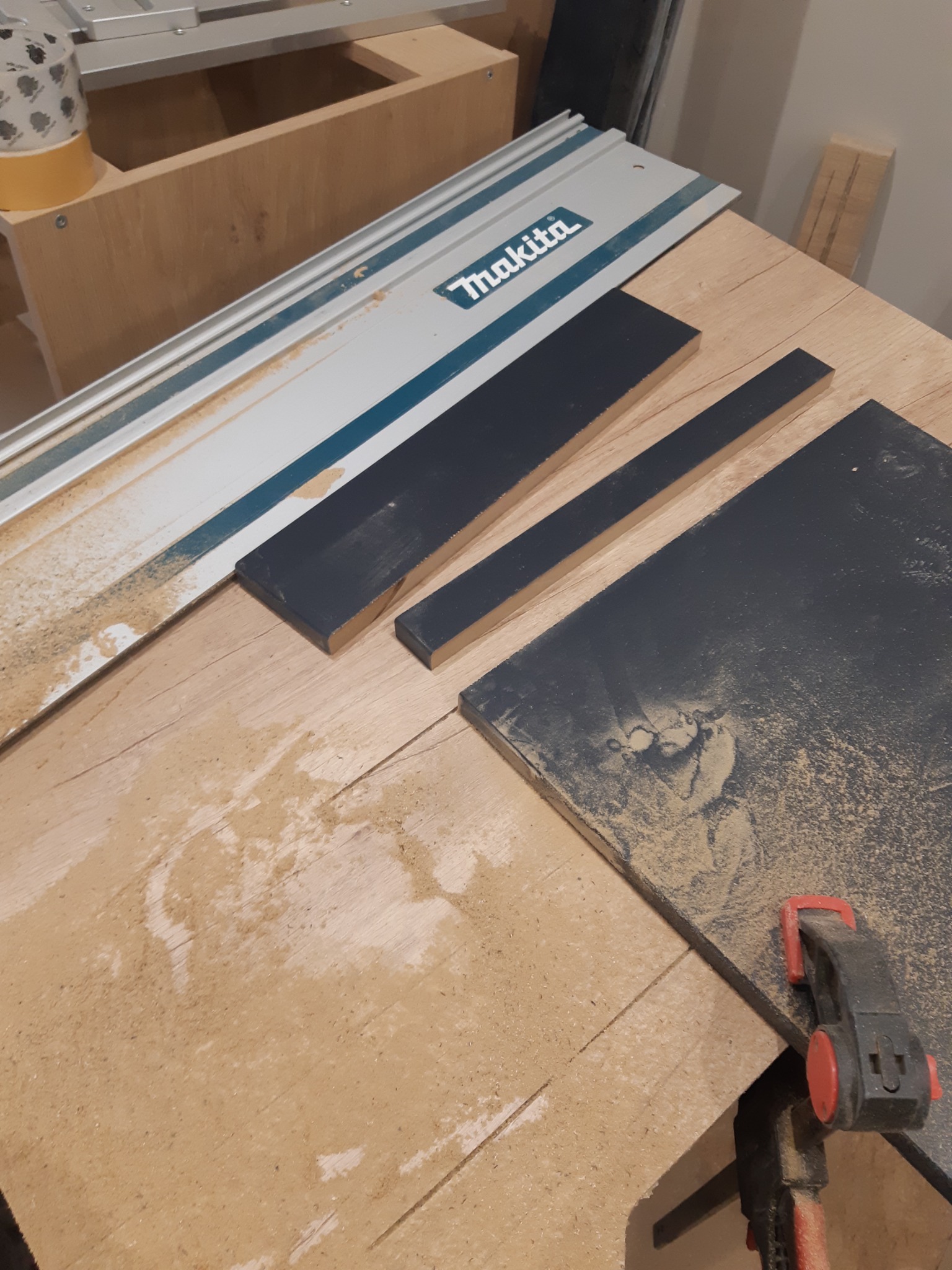Przygotowane, pomalowane elementy drewniane leżące na stole warsztatowym z szyną prowadzącą Makita i śladami pyłu po obróbce, zabezpieczone ściskiem stolarskim.