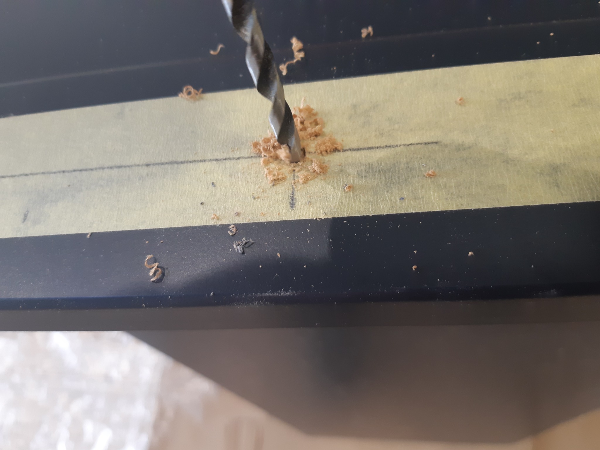 Wiertło wiercące otwór w czarnej płycie, zabezpieczonej taśmą malarską z zaznaczonym punktem wiercenia. Wióry wokół wiertła.