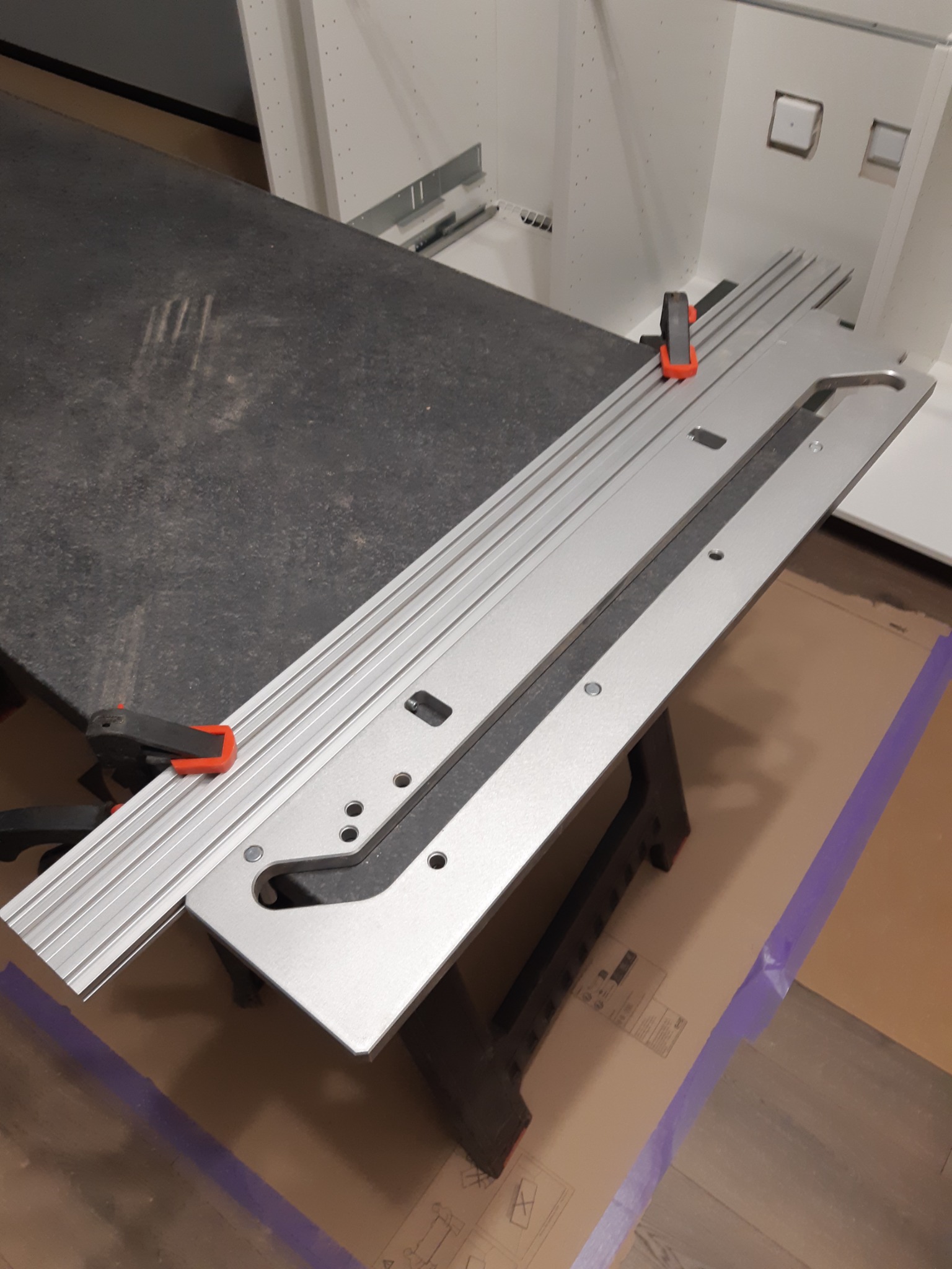 Szablon do montażu mebli kuchennych, przymocowany klamrami do blatu. Widoczne elementy szafek w tle. Przygotowanie do instalacji.