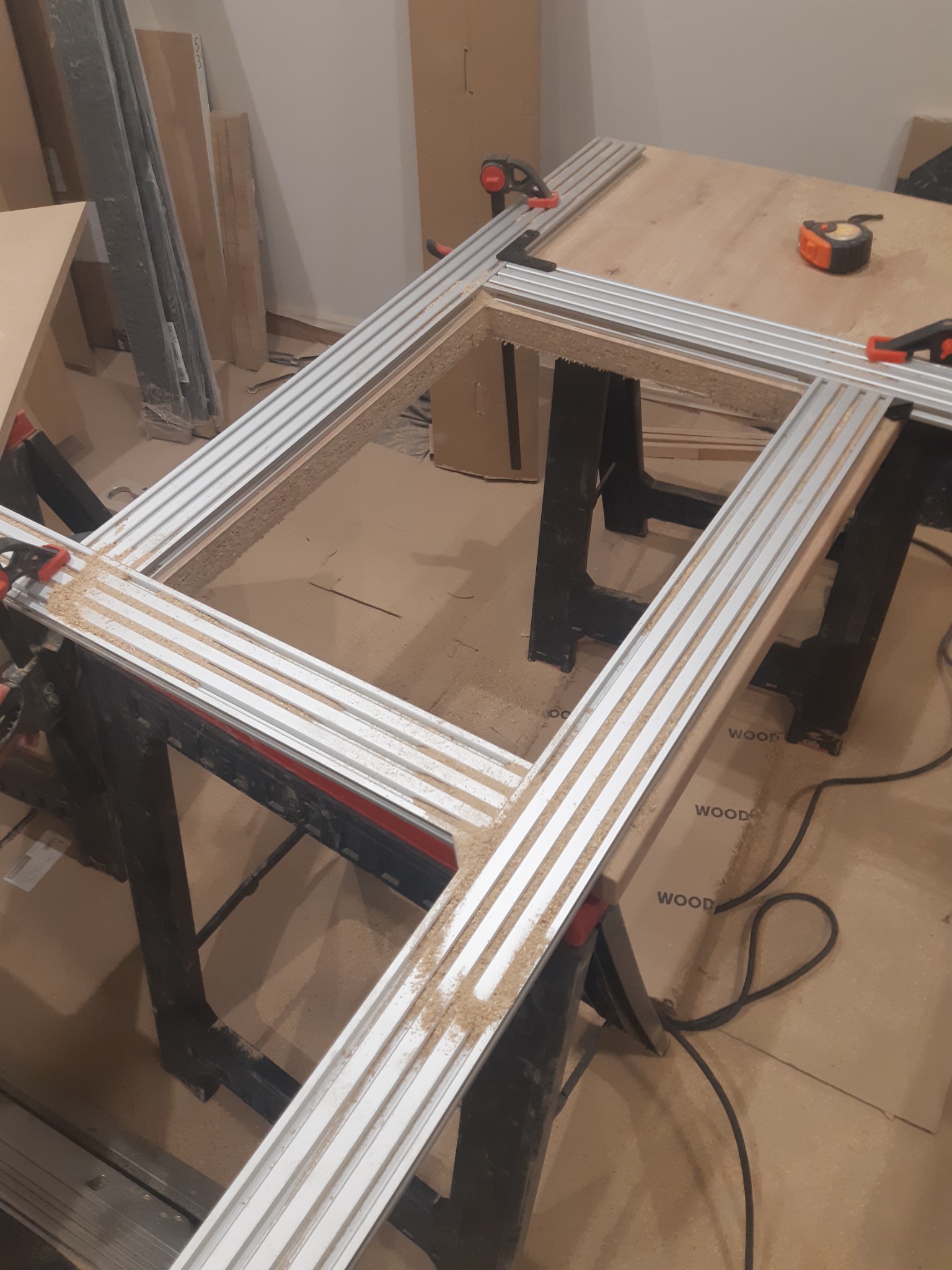 Rama meblowa z profili aluminiowych, zabezpieczona zaciskami, leżąca na kozłach. Widoczne ślady obróbki drewna i miara zwijana. Przygotowanie do montażu.