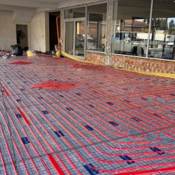 6/4 CALA - Czerwone rury ogrzewania podłogowego ułożone na siatce w surowym wnętrzu budynku z dużymi oknami, widok z góry, etap instalacji przed wylewką.