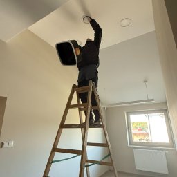 6/4 CALA - Instalator na drabinie montuje okrągły nawiewnik sufitowy, trzymając urządzenie pomiarowe Testo. Widoczne okno i grzejnik w jasnym pomieszczeniu.