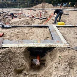 6/4 CALA - Instalacja rur w piasku na budowie, widoczne fundamenty i pracownicy. Pomarańczowe rury częściowo zakopane, żółta skrzynka narzędziowa w tle. Prace ziemne w toku.