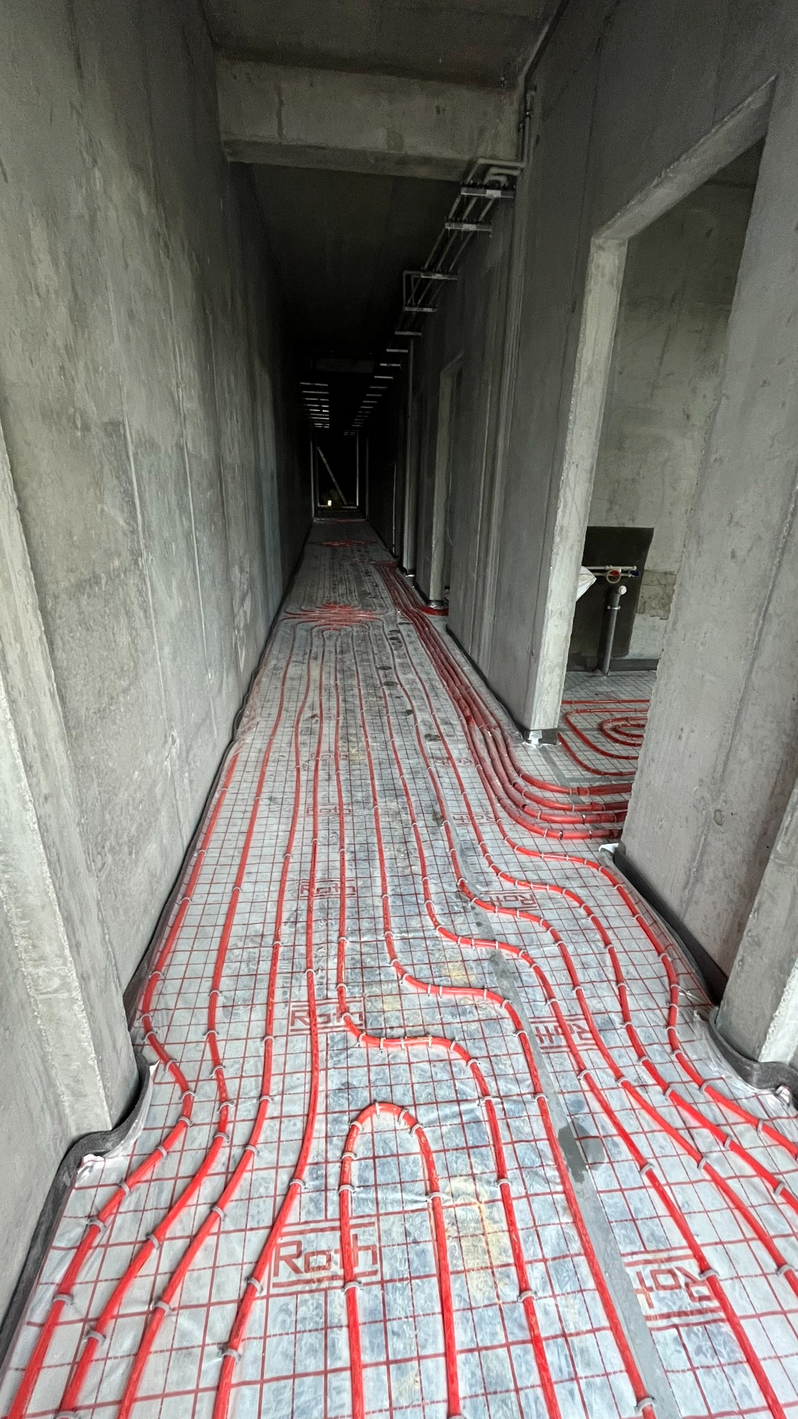 Czerwone rury ogrzewania podłogowego ułożone na szarej macie w surowym wnętrzu korytarza z betonowymi ścianami. Widok w perspektywie w głąb korytarza.