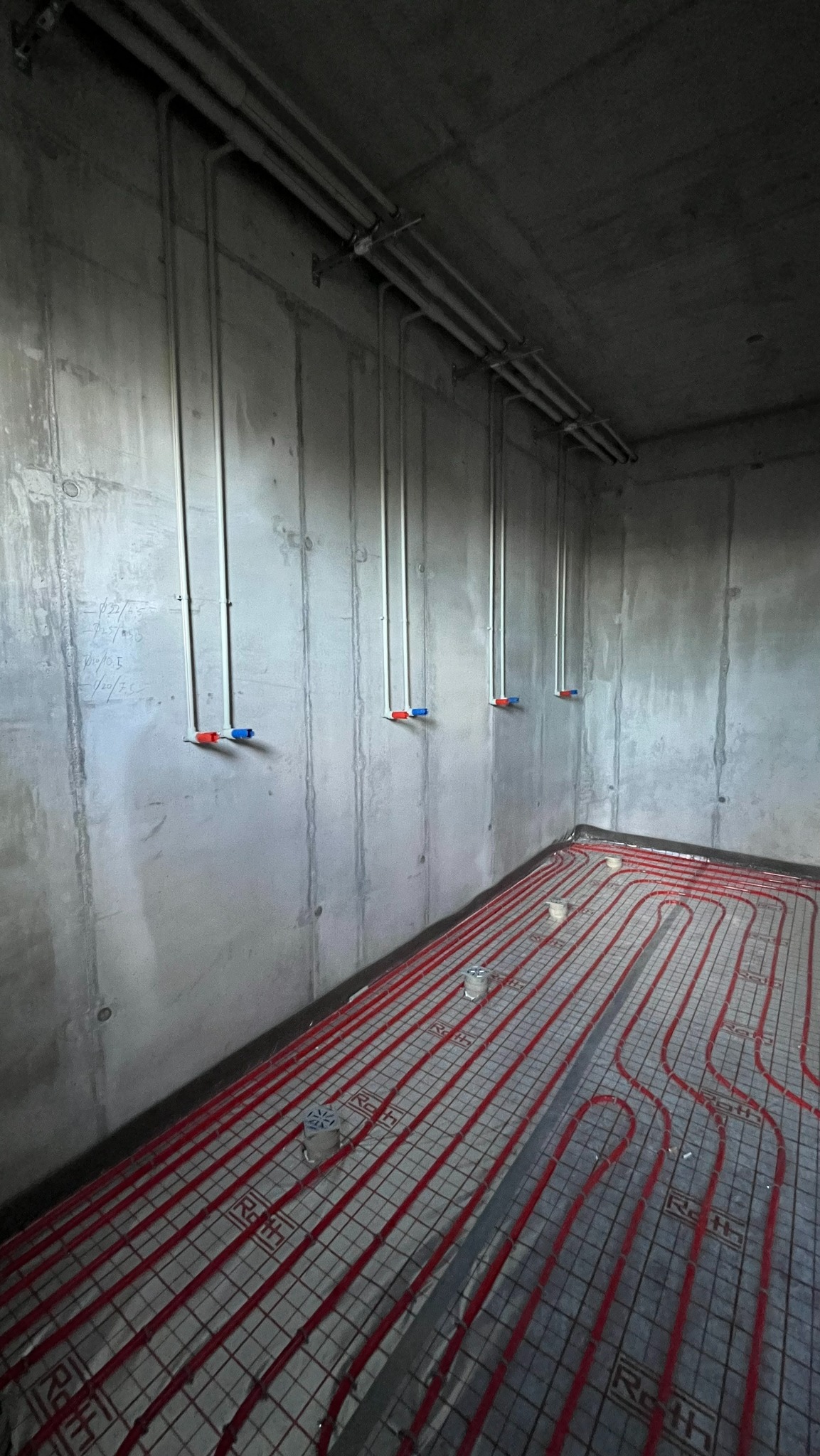 Szare rury pionowe z czerwono-niebieskimi końcówkami na ścianie nad czerwoną instalacją ogrzewania podłogowego Roth w surowym wnętrzu. Widok z góry.