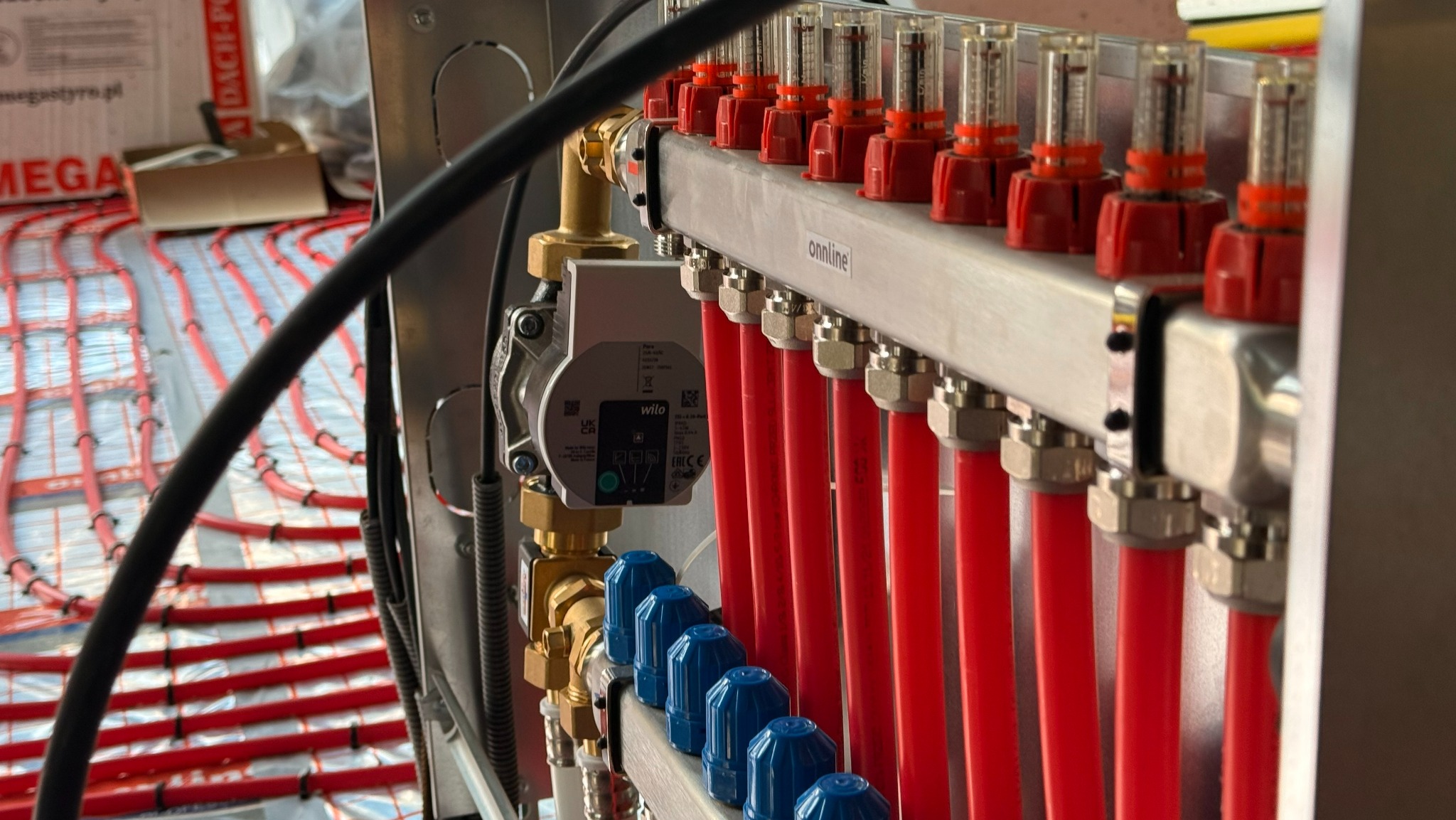 Rozdzielacz ogrzewania podłogowego z czerwonymi rurami, widoczna pompa obiegowa Wilo i elementy regulacyjne. W tle ułożone rury ogrzewania podłogowego na izolacji.