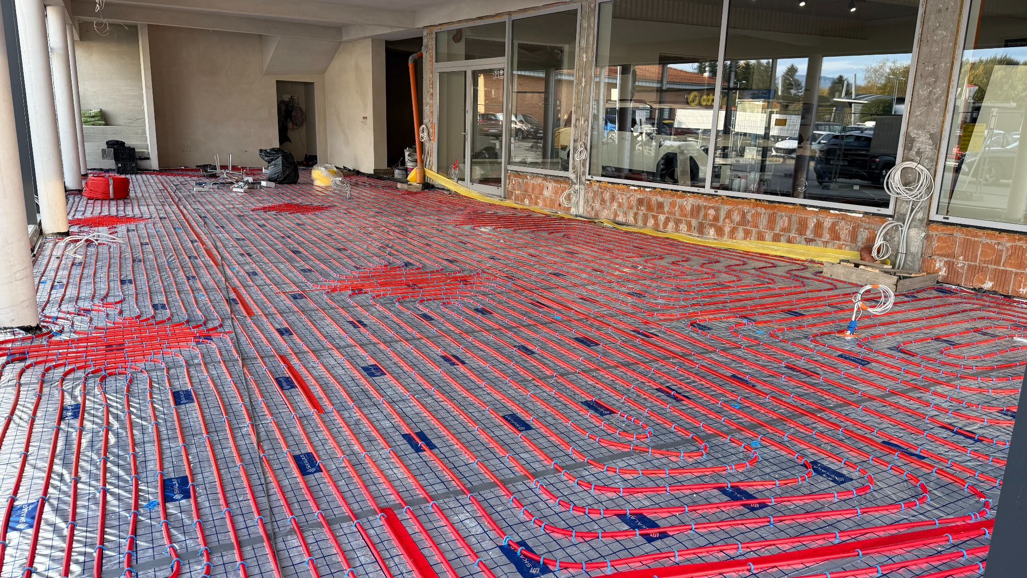 Czerwone rury ogrzewania podłogowego ułożone na siatce w surowym wnętrzu budynku z dużymi oknami, widok z góry, etap instalacji przed wylewką.